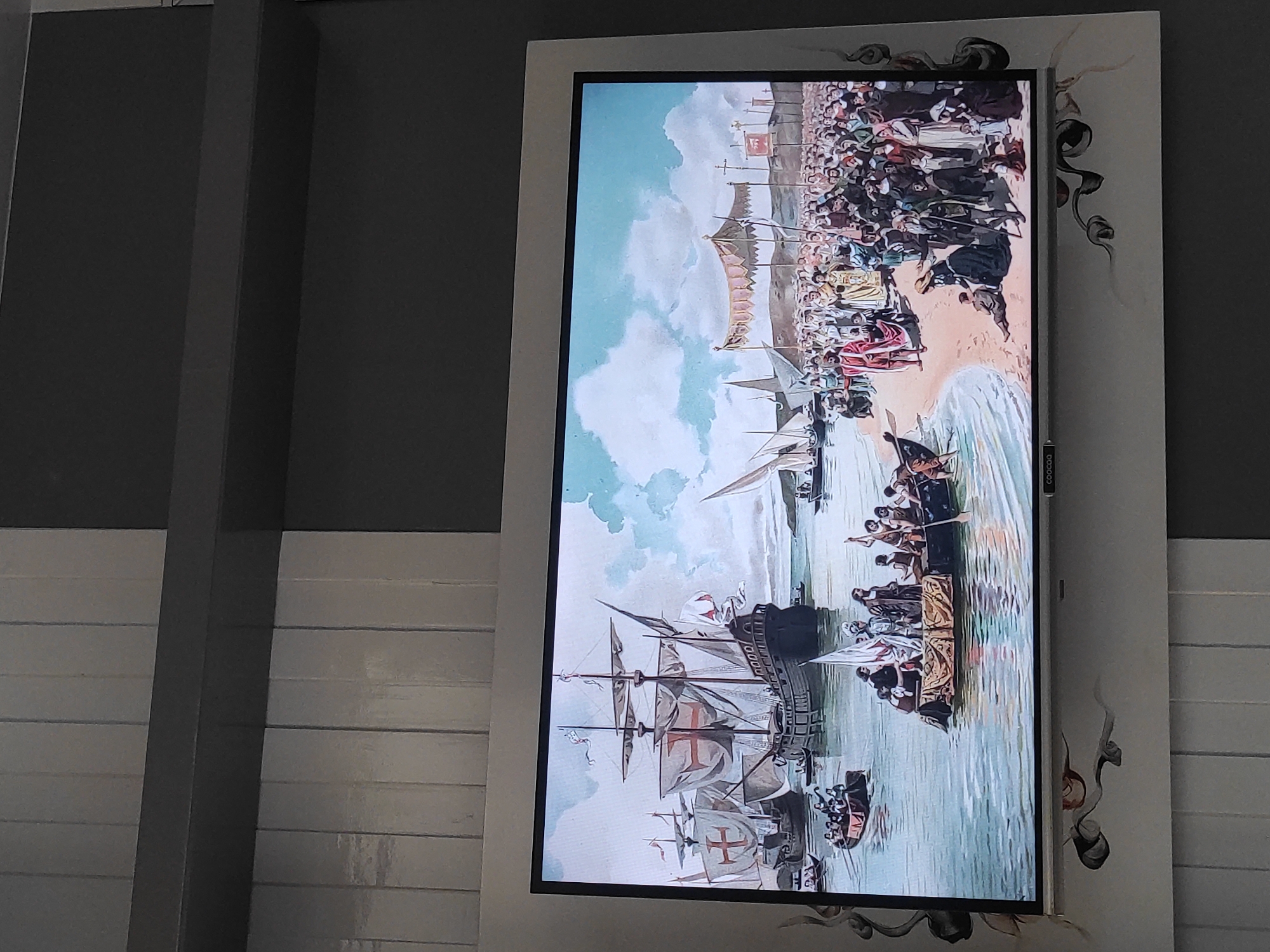 The image displays a framed digital screen showing a historical painting of a maritime scene. The painting depicts several large sailing ships with red crosses on their sails in a harbor. In the foreground, a group of people are gathered on a beach, some disembarking from a smaller boat. Further out in the water, more boats and ships are visible. The sky is cloudy with a bright blue expanse. The digital screen is mounted on a wall with a decorative border featuring floral motifs. The background wall has a pattern of vertical grey panels. The bottom edge of the screen shows a logo that appears to read "COOCOQ". The context provided indicates this scene is displayed in Sumedang, Indonesia, though the painting itself does not contain any elements that specifically identify it as being related to Sumedang.
