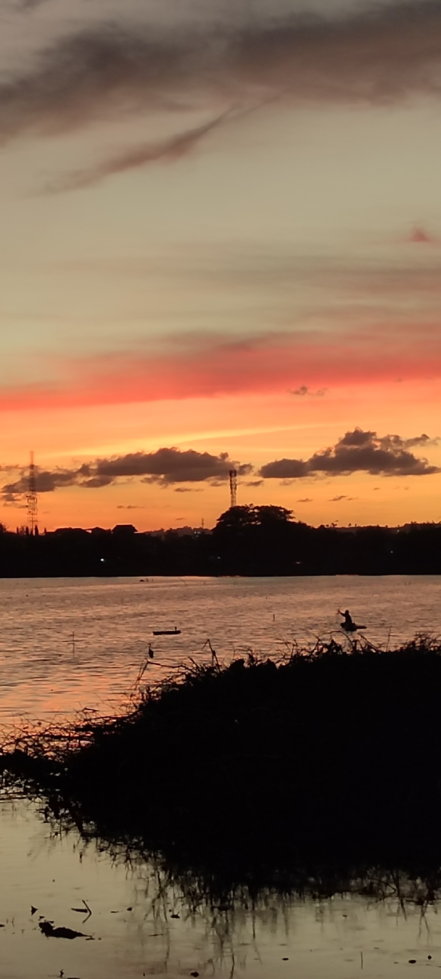This is a vertical, low-angle, wide shot of a sunset over a body of water, likely a lake or reservoir, at the edge of Lhokseumawe, Indonesia. The sky displays a gradient of colors, transitioning from a pale yellow at the top to a vibrant orange and red band across the middle, with dark grey clouds scattered throughout. The horizon line is silhouetted by the dark shapes of trees, buildings, and at least two tall transmission towers. On the water, there are several small, dark objects, possibly boats, and a single bird perched on a thin branch in the foreground. The foreground is dominated by the dark, silhouetted outline of dense vegetation and a thin branch with a bird on it, casting reflections on the water. The water itself is calm, with ripples reflecting the colors of the sunset.