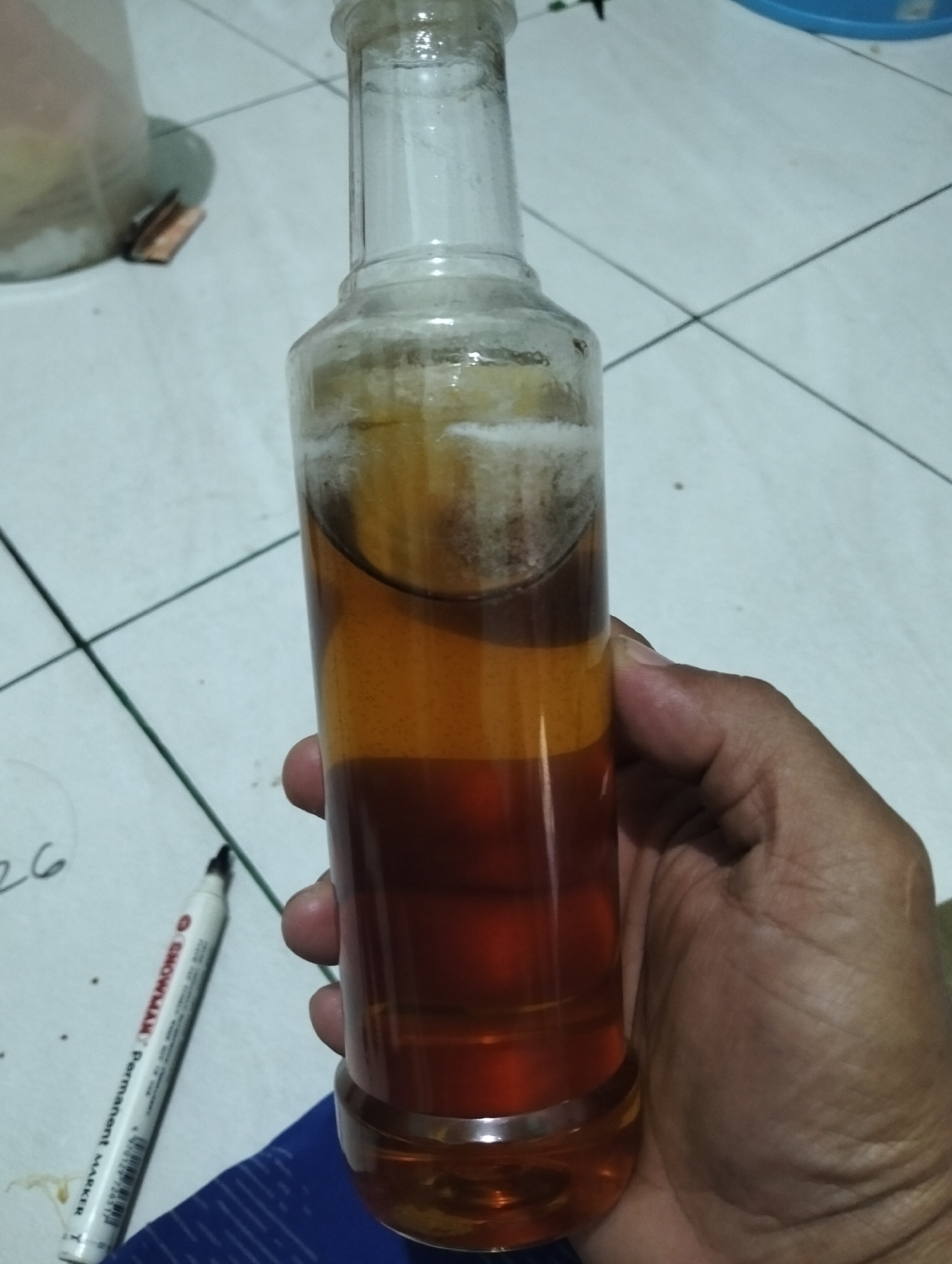 A hand holds a clear glass bottle filled with a liquid that has separated into distinct layers of amber and darker red. The bottle is tall and cylindrical with a narrow neck and a wider base. A white marker with red text, identified as "SNOWMAN Permanent MARKER," lies on a blue and white patterned surface to the left of the bottle. The background consists of a tiled floor with visible grout lines, suggesting an indoor setting. The overall impression is of a scientific or domestic setting, possibly in Paciran, Indonesia, given the context.
