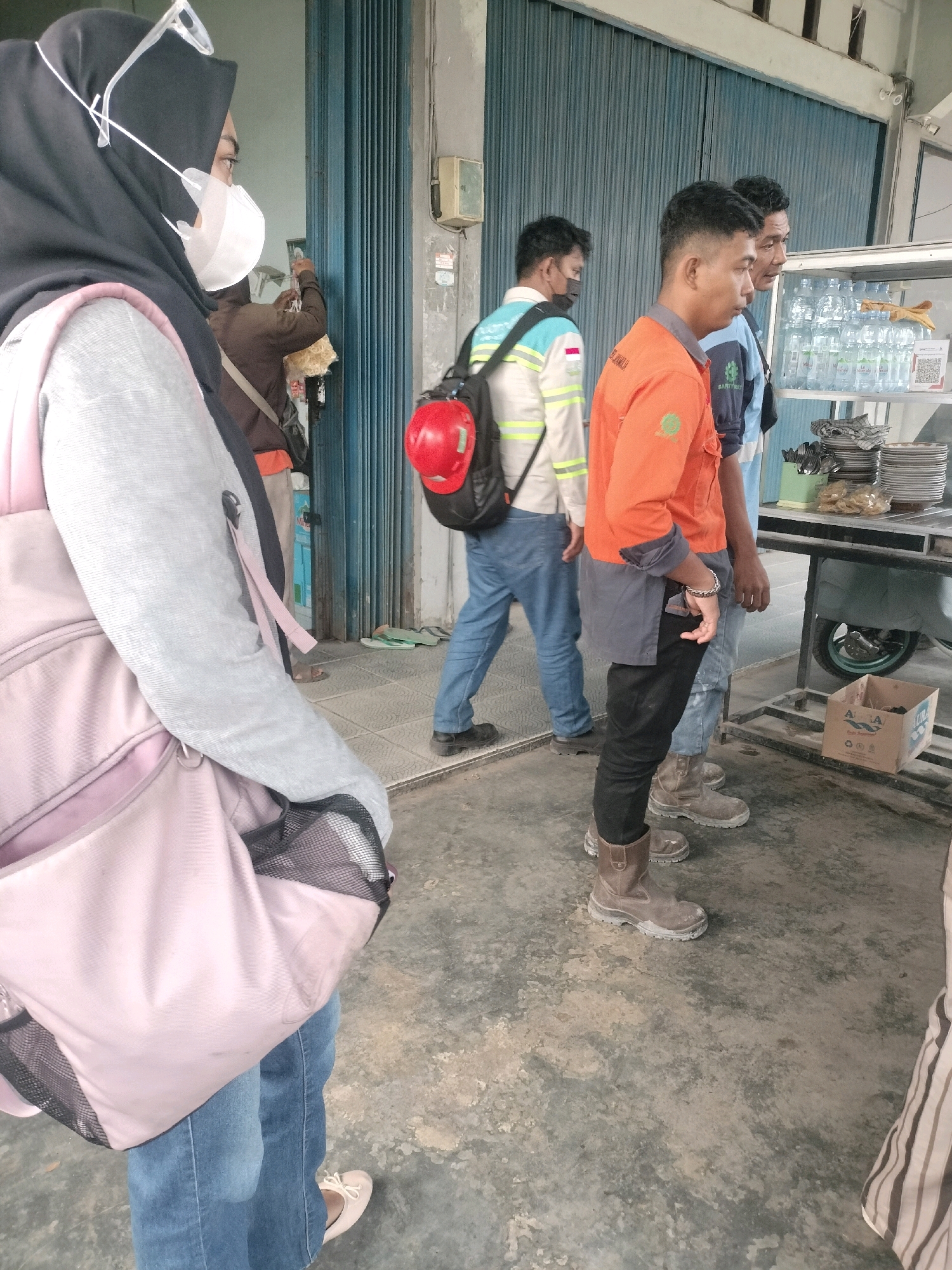 In Maburai, Indonesia, an outdoor scene unfolds with a blue corrugated metal door in the background. Several individuals are present. A person in the foreground, wearing a black hijab and a light grey jacket over blue jeans, carries a light pink backpack. To their right, another individual in a brown jacket is visible. Further back, three men are seen. One is wearing a white and light blue jacket with reflective yellow stripes and a red hard hat. Another man is in an orange work shirt with grey accents over blue jeans, also wearing work boots. A third man to his right wears a blue shirt. A metal food cart with stacked plates, bottles of water, and snacks is positioned on the right side of the frame. A cardboard box is on the ground near the cart. The ground is a worn, concrete-like surface.