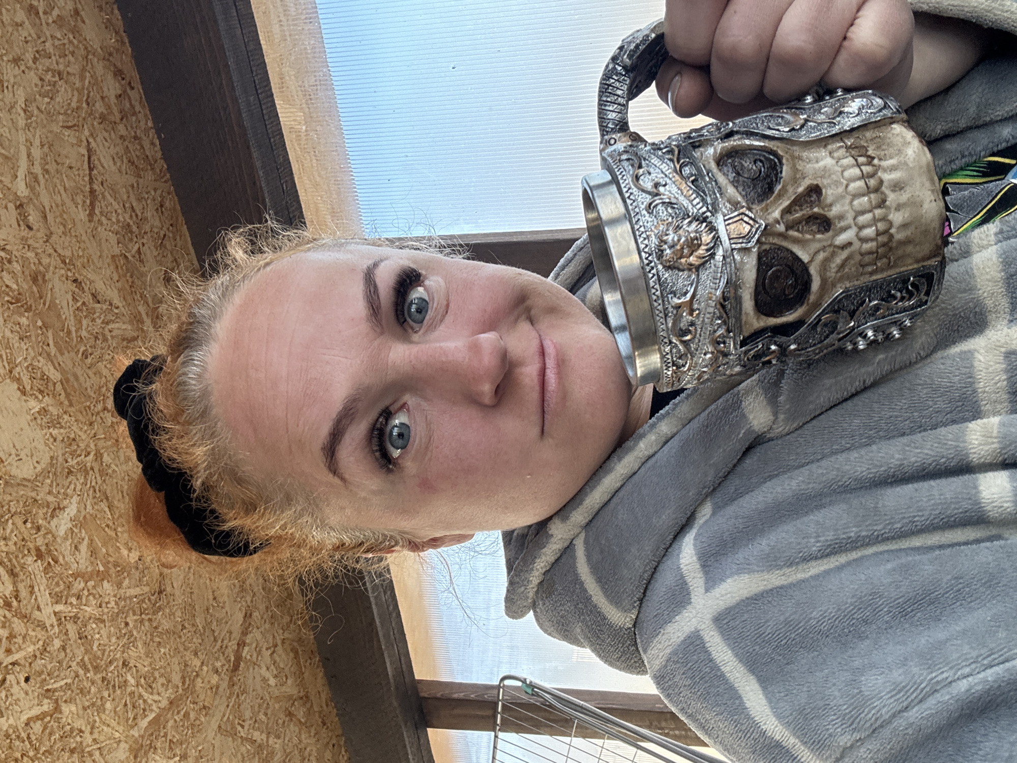 A woman with fair skin and blue eyes is holding a large, ornate skull-shaped mug. She is wearing a gray robe with a white plaid pattern. Her hair is pulled back into a bun. The background includes a wooden wall and a transparent ceiling. The location is Chaikovsky, Russia.