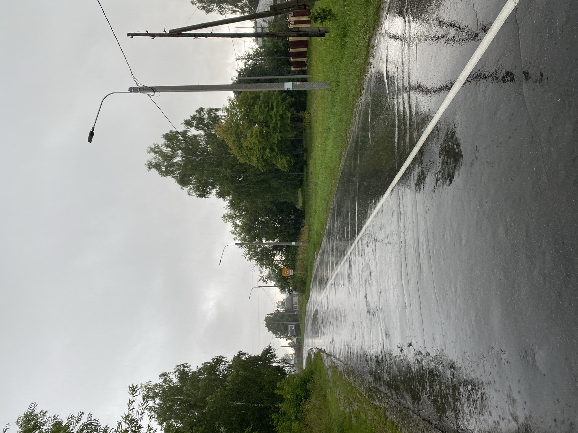 A wet asphalt road stretches into the distance under a cloudy, overcast sky, with visible raindrops in the air. The road has a white dividing line. Green trees and grass line both sides of the road. On the right side, there are utility poles and a wooden fence. In the background, there are some houses and a yellow road sign depicting children. The scene appears to be a rural or suburban area during a rain shower.