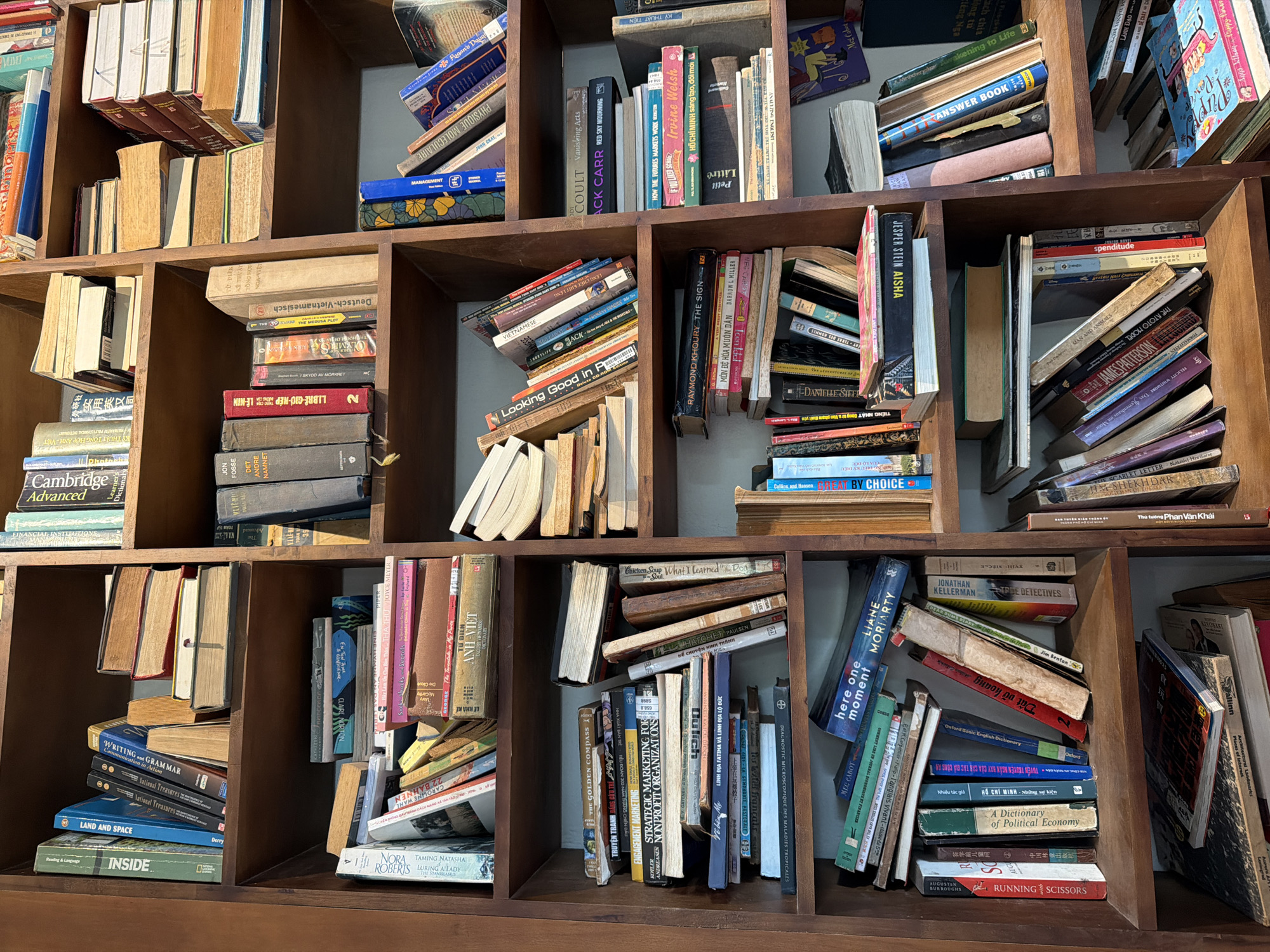 This image shows a close-up view of wooden bookshelves filled with numerous books in a room. The books are of various sizes, colors, and states of condition, with some appearing well-worn and others newer. They are arranged both vertically and horizontally on the shelves, with some leaning against each other. The lighting suggests an indoor environment, possibly a library, bookstore, or a private collection. The presence of Vietnamese titles and languages on some of the book spines indicates that this scene is located in Vietnam. Given the detail of the books and their arrangement, the establishment appears to be a well-stocked library or bookstore, potentially in a city like Da Nang, Vietnam, which is known for its cultural attractions and growing number of bookstores. There are no people visible in the frame, and the focus is entirely on the books and the shelving unit. The scene is static and does not depict any actions or interactions.