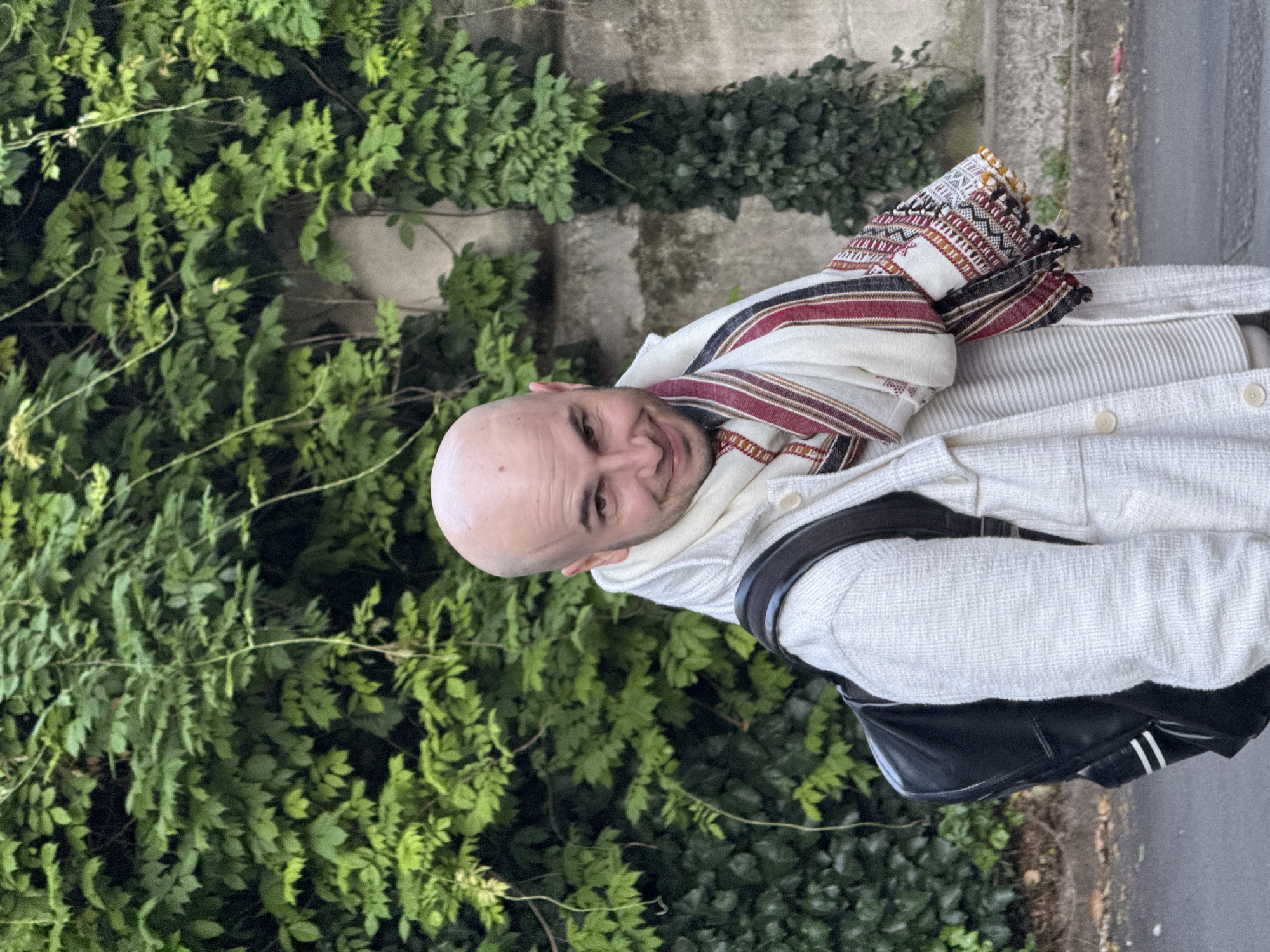 The image shows a bald man with a slight smile, wearing a cream-colored jacket, a patterned scarf, and a black backpack. He is standing outdoors in front of a wall covered in green ivy and foliage. To the right of the wall appears to be a paved surface. The image is likely taken in an outdoor setting, potentially within a city environment. It is not possible to determine whether this picture was taken in Milan, Italy without additional information.