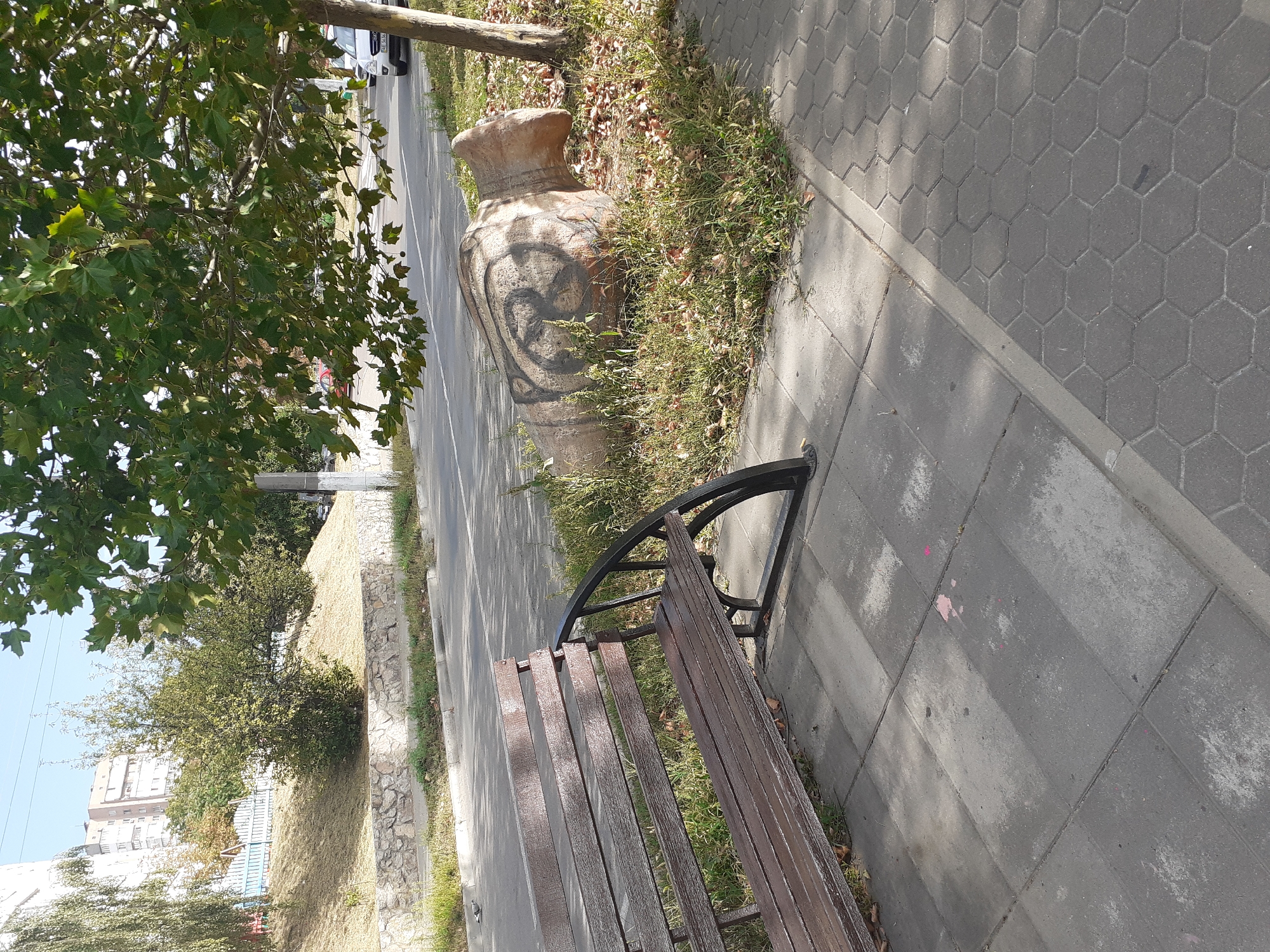 The image depicts a street scene in Obukhiv, Ukraine. In the foreground, a weathered wooden bench with a black metal frame sits on a grey tiled sidewalk. To the right of the bench, a large, concrete object resembling a vase or urn lies on the ground, partially overgrown with grass and fallen leaves. This object has a crude, dark spray-painted design on its side. Behind the bench and the concrete object, a road is visible with some parked cars and street markings. A slope with dry grass and a low stone wall borders the road. Further in the background, a multi-story building and trees are visible under a clear sky. The overall impression is a somewhat neglected urban environment.