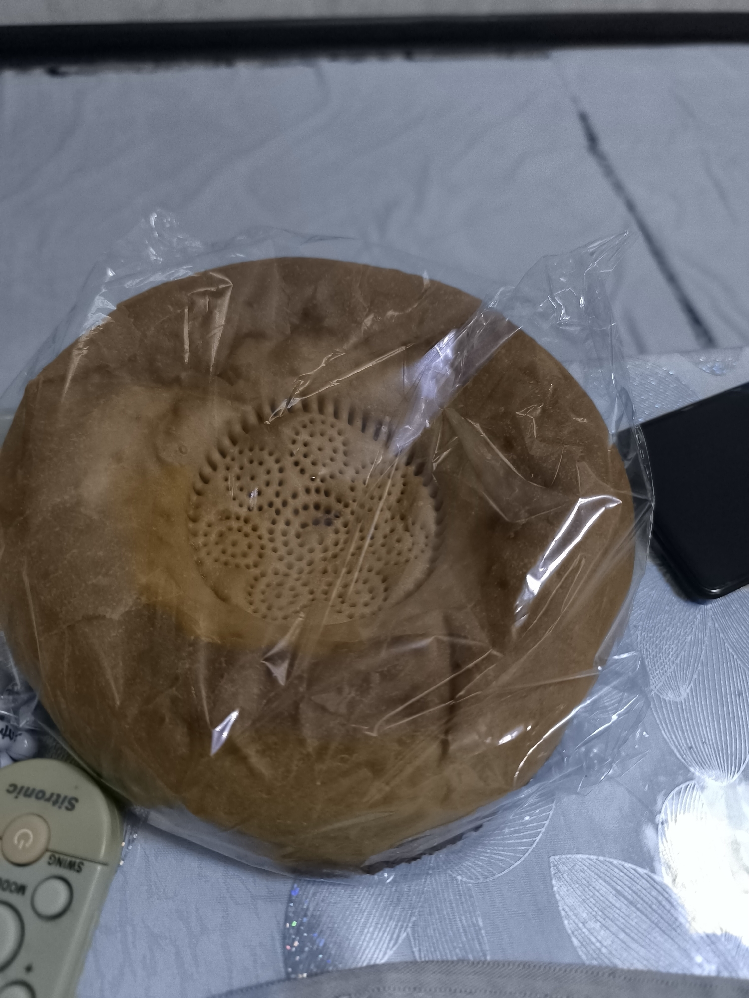 A round, flatbread, likely a lepeshka, is wrapped in clear plastic and lies on a blue and white patterned fabric surface, possibly a bedspread. The bread has a golden-brown crust and a textured center with many small indentations. To the right of the bread, a black smartphone is visible. In the lower left corner, a light-colored remote control with buttons and lettering, including "Sitronic" and "SWING," is present. The scene appears to be indoors, in a domestic setting. The image is taken from a slightly elevated angle. The specific location in Kokand, Uzbekistan, cannot be determined from the visual information alone.