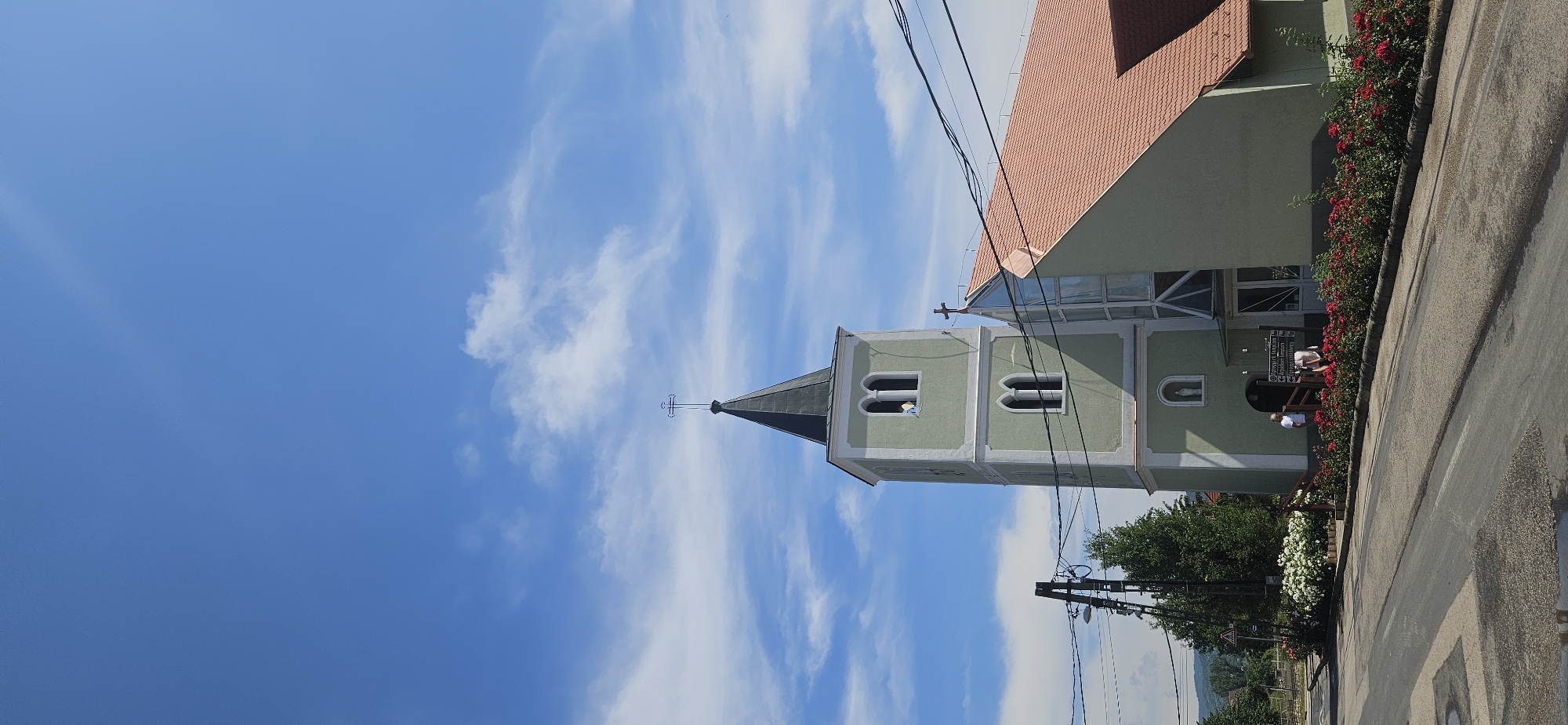 The image shows the exterior of a church in Hévíz, Hungary. The church has a tall, light green tower with a dark pointed roof topped by a cross. The tower has arched windows and a small niche with a statue of a figure. Adjacent to the tower is the main body of the church, also light green, with a terracotta-tiled roof. A large modern window structure is visible on the side of the church. In front of the church, there is a road with some power lines running across the frame. A line of red roses is planted along the edge of the road, with a cluster of white flowers and greenery further back. Two individuals are standing near the entrance of the church. The sky above is a bright blue with scattered white clouds.