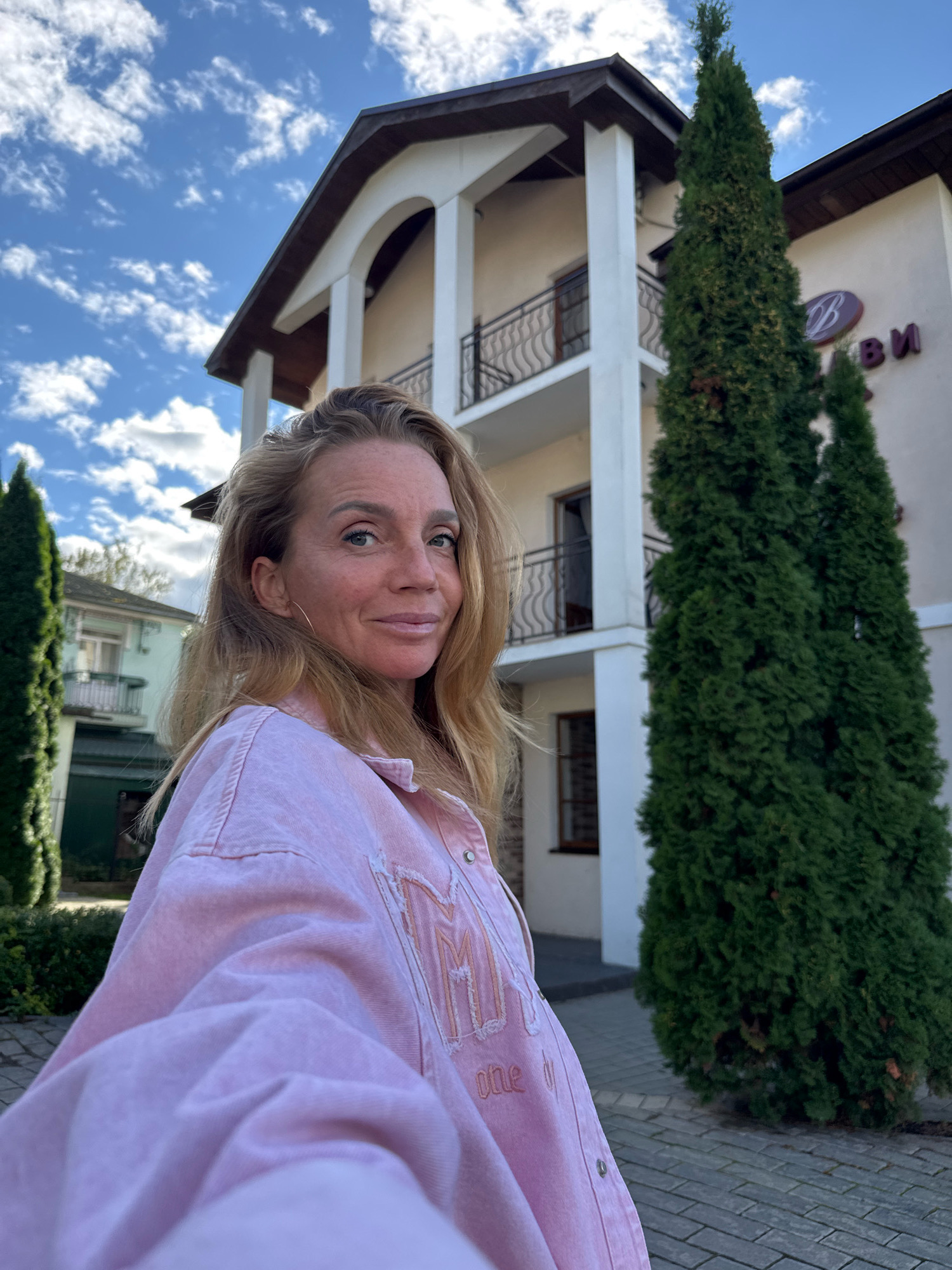 A woman with blonde hair and fair skin is standing in front of a building in Staraya Russa, Russia. She is wearing a pink denim jacket. The building appears to be a hotel or guesthouse, with a white facade, arched windows, and balconies. Two tall, green cypress trees stand in front of the building, framing the woman. The sky is blue with scattered white clouds. The ground is paved with grey bricks.