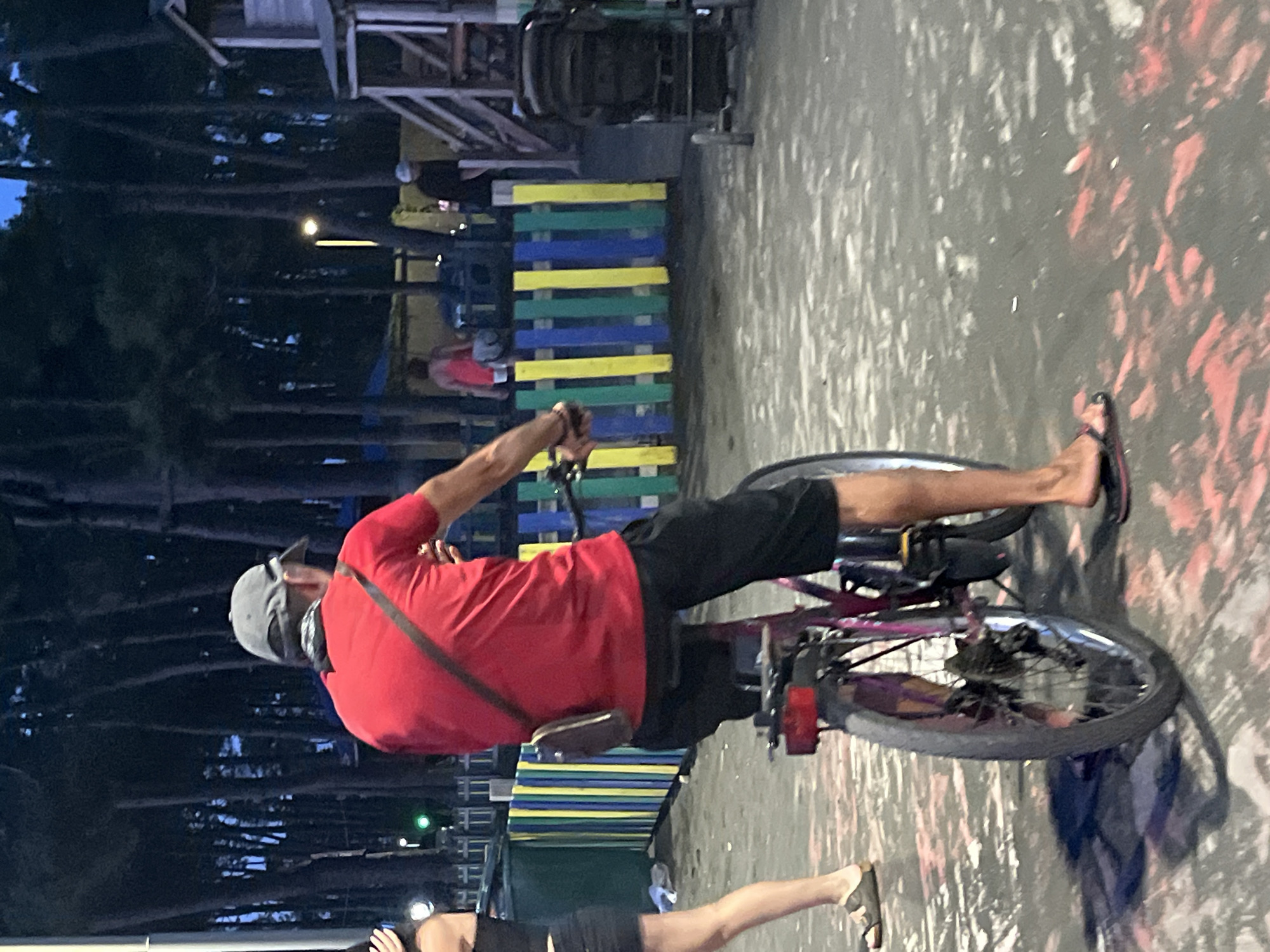 In Ureki, Georgia, at dusk, a man wearing a red t-shirt, black shorts, and a grey cap rides a bicycle. He has a small brown bag slung across his chest. The scene takes place on a paved area with red and white markings. Behind him, a colorful wooden fence, painted in vertical stripes of yellow, blue, and green, separates the paved area from a densely wooded area of tall, dark trees. Further in the background, a wooden structure, possibly a playground, is visible, along with other individuals and a stroller. A green light is illuminated on the left side of the image.