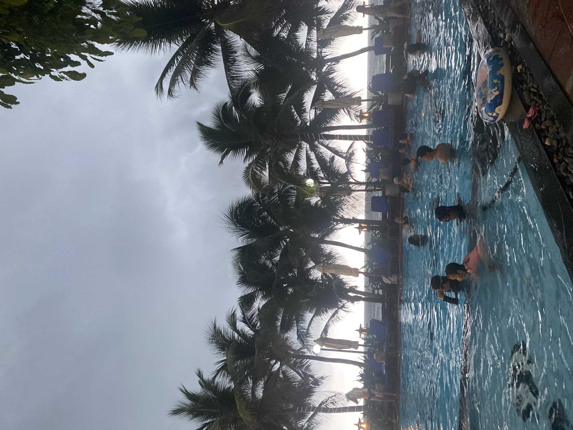 The image captures a vibrant scene at an outdoor swimming pool, likely part of a tropical resort in Phu Thuy Ward, Vietnam. The perspective frames the blue swimming pool in the foreground and midground, with a dense line of tall palm trees silhouetted against the sky and the distant sea.
Numerous people, including children and adults, are enjoying themselves in the pool. They are seen swimming, wading, and relaxing in the water. On the dark, tiled edge of the pool in the foreground, a blue and yellow inflatable swimming ring adorned with star patterns is visible, accompanied by a pair of pink flip-flops.
Behind the pool, the prominent feature is a row of majestic palm trees, their fronds dark against the brighter background. Beyond the palm trees, a distinct, bright line indicates the horizon where the sea meets the sky. The sky above is overcast with soft, grey clouds, suggesting either late afternoon or early morning light, which creates the silhouetted effect on the palm trees. Among the palm trees, several blue lounge chairs with white umbrellas are arranged, indicating a relaxing poolside area overlooking the ocean. A few outdoor lights or lanterns are also visible between the chairs. The scene conveys a sense of leisure and enjoyment in a beautiful coastal setting. No discernible text is visible on any objects within the image.