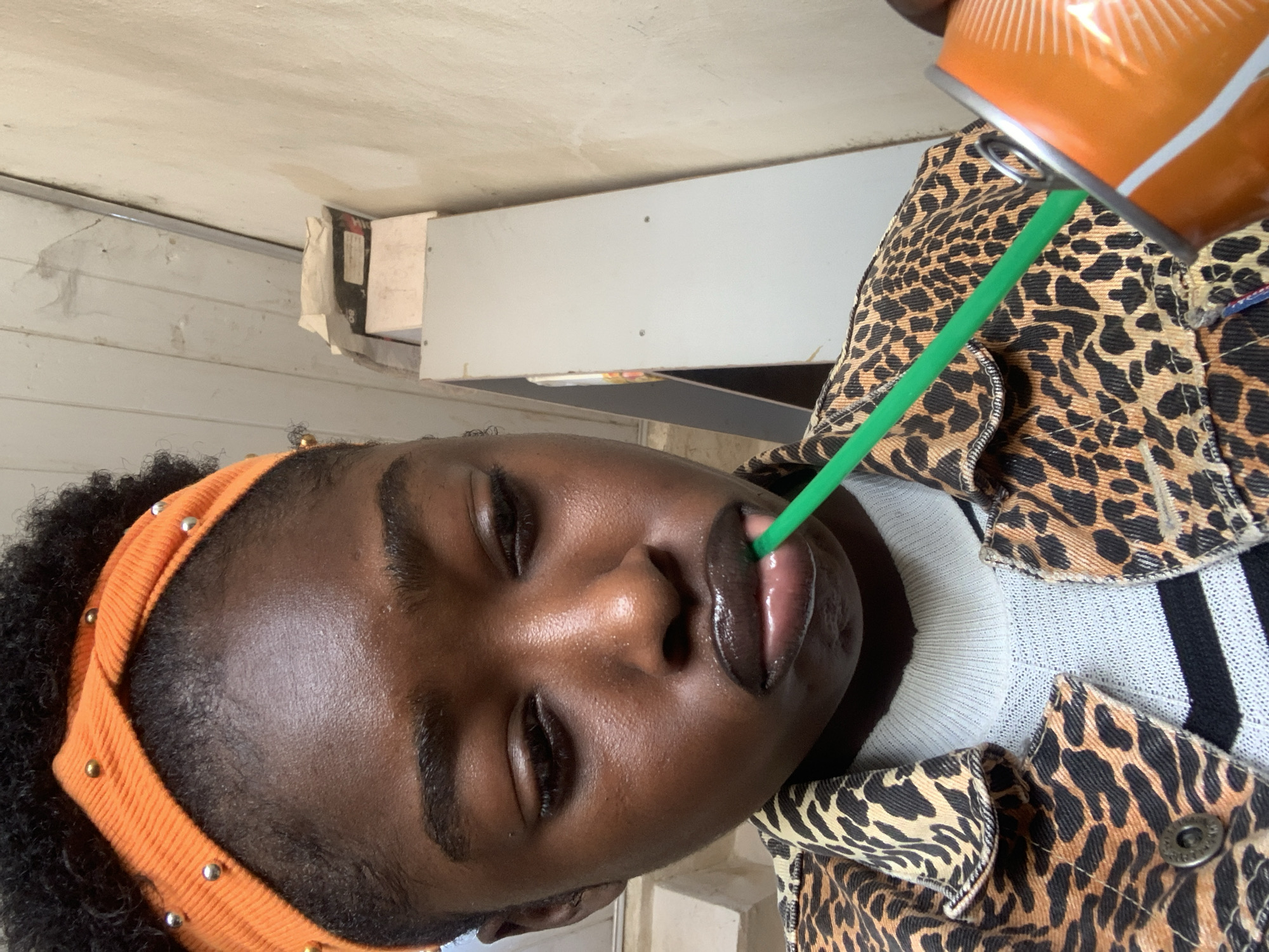 A close-up, medium shot captures a young Black person indoors, their face angled down towards a bright orange and white can with a silver pull tab, held in their right hand. A vibrant green straw extends from the can into the person's mouth, where their lips are slightly parted. The person wears an orange, ruched headband adorned with small silver and gold beads. Their attire includes a leopard print jacket over a white and black striped top. The background shows a textured, off-white wall with wooden paneling and shelves stacked with various items, suggesting an interior space possibly in a residential area of New Stadium, Nigeria. The lighting is overhead, highlighting the person's glossy lips and the smooth texture of their skin.