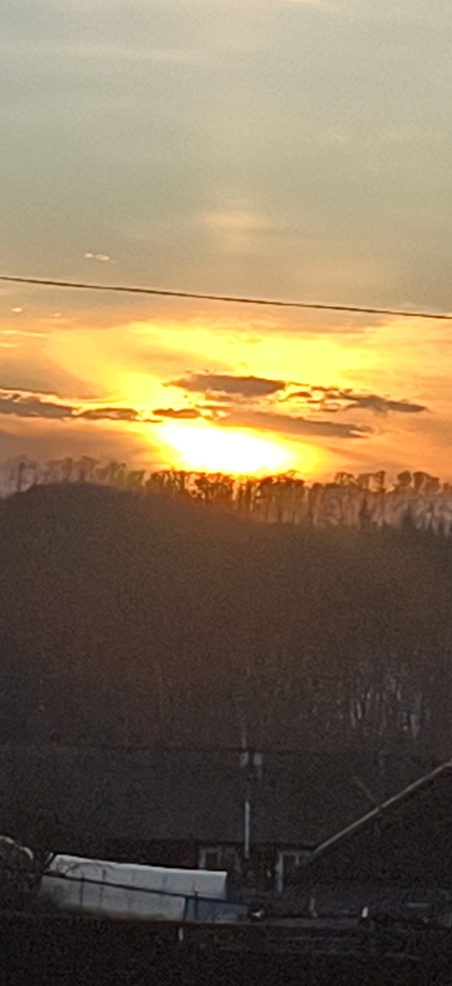 A scenic view captures a bright sunrise or sunset, casting a warm glow across the sky and landscape. Above the horizon, layers of clouds are illuminated in hues of yellow and orange, creating a vibrant and dynamic atmosphere. An overhead power line, running horizontally across the top third of the image. A dark, silhouetted ridge runs across the midground, with the sun or light source positioned behind it, creating a stark contrast. This ridge appears to be a dense tree line. In the foreground, details are difficult to discern due to darkness, but there appears to be a low structure or building and angled piping. Overall, the scene conveys a peaceful, natural setting. I don’t have specific information on the location, Фоки, Russia, to make any accurate comparisons.