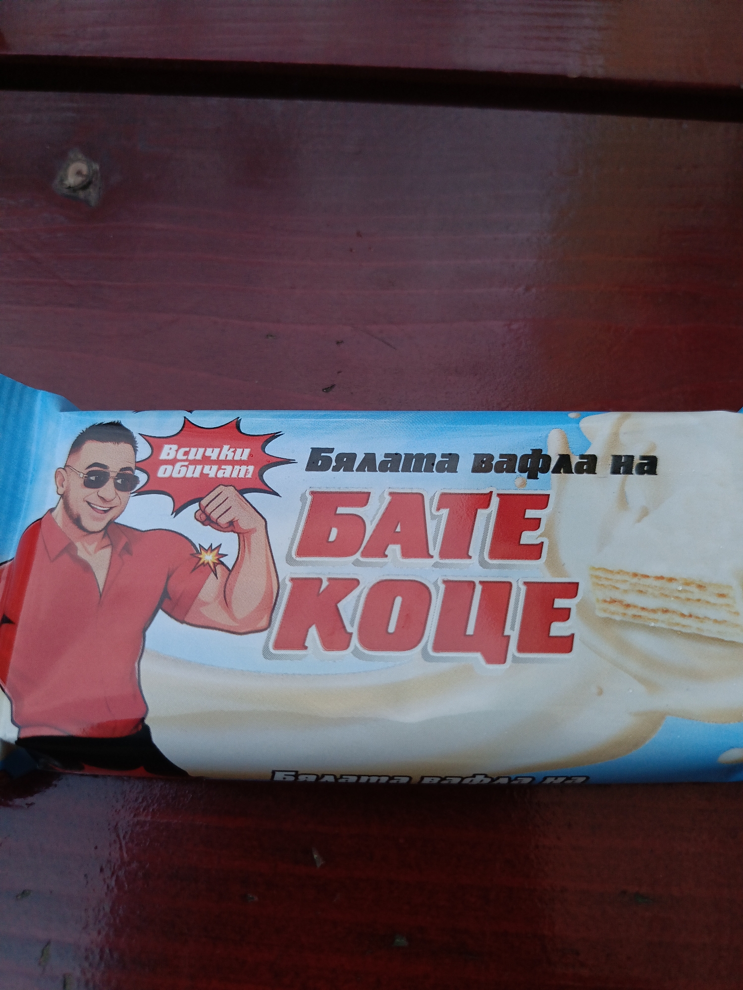 The image shows a close-up of a product wrapper, likely for a candy bar or snack, lying on a dark red wooden surface. The wrapper features a cartoon illustration of a muscular man with sunglasses and a red shirt, flexing his bicep. To the right of the illustration, large red text reads "BATE KOЦE" (Bate Koce), with the words "Бялата вафла на" (The white waffle of) above it. Smaller text within a starburst shape near the man's head says "Всички обичам" (Everyone loves). A portion of the product, appearing to be a layered waffle with white filling, is visible on the right side of the wrapper. The background is a blurred dark red wooden texture, possibly a table or bench. There are no identifiable landmarks or indications of the city of Varna, Bulgaria in this image.