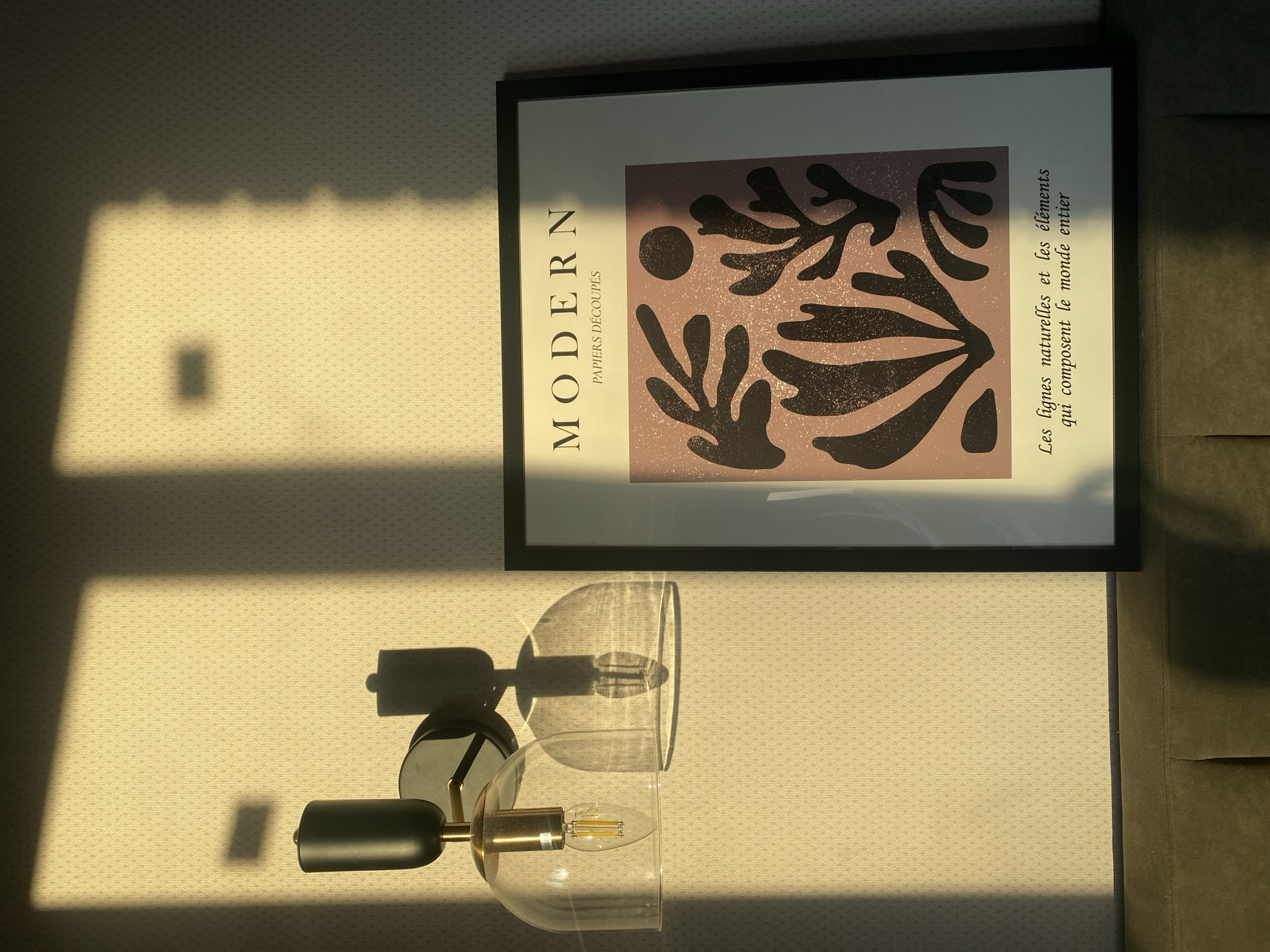 The image shows a wall with a black-framed art print, and a wall-mounted lamp. The print displays an abstract design in mauve and black on a white background. The words "MODERN PAPIERS DÉCOUPÉS" are printed vertically along the left side of the image, with the phrase "Les lignes naturelles et les éléments qui composent le monde entier" is printed vertically along the right side. Below the print is a black and gold wall-mounted lamp with a transparent glass shade that encases a light bulb. Sunlight casts shadows across the wall, creating a pattern of light and dark. The wall appears to be covered in a textured wallpaper. The city and specific location are not discernable.