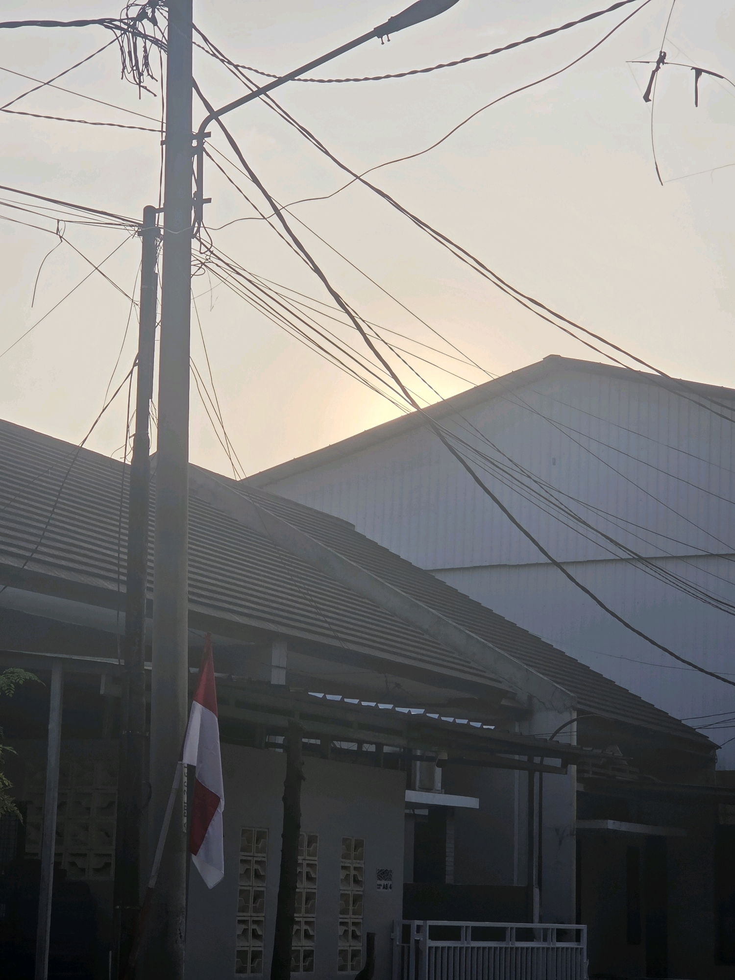 The image depicts a street scene in Bojongsoang, Indonesia, under a hazy sky. A prominent utility pole stands on the left side, adorned with numerous tangled electrical wires that crisscross the frame. A small Indonesian flag, red and white, hangs from the pole. Behind the pole, two houses with tiled roofs are visible. The house on the left has a grey facade with patterned windows, while the house on the right is a larger, white structure. The sun is partially obscured by the haze, casting a soft glow in the upper right portion of the image. The overall impression is one of a residential area with dense infrastructure.