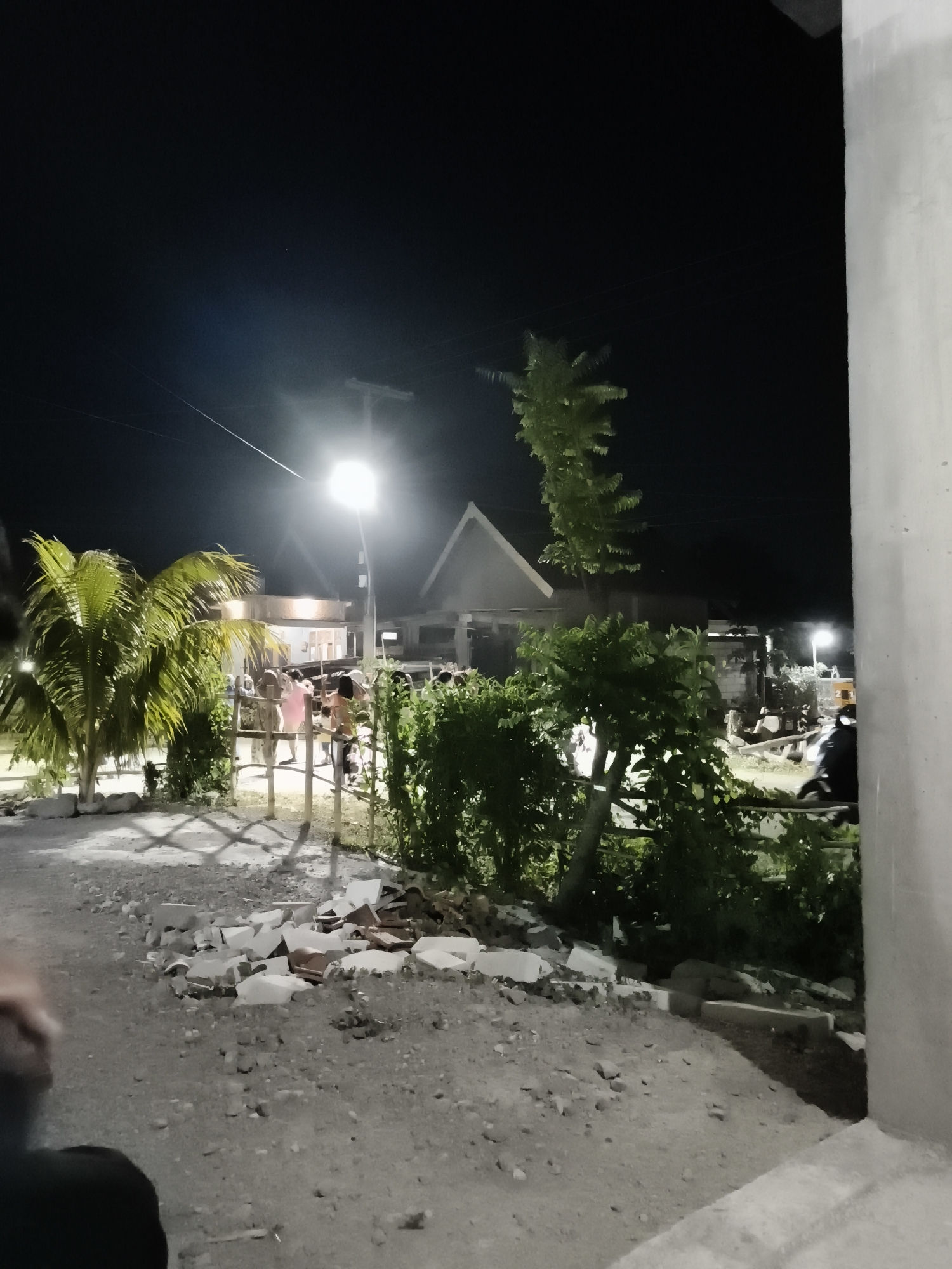 The image captures a nighttime scene in Pandean, Indonesia, likely a public area or pathway. A bright streetlamp illuminates the surroundings, casting shadows and highlighting a pile of broken concrete and tiles in the foreground. To the left, a palm tree stands, its fronds catching the light. In the background, a group of people are gathered near a fence and some buildings, their activities unclear due to the low light and distance. The environment appears somewhat unkempt, with scattered debris and vegetation. The overall impression is of a functional, albeit somewhat disheveled, community space at night.