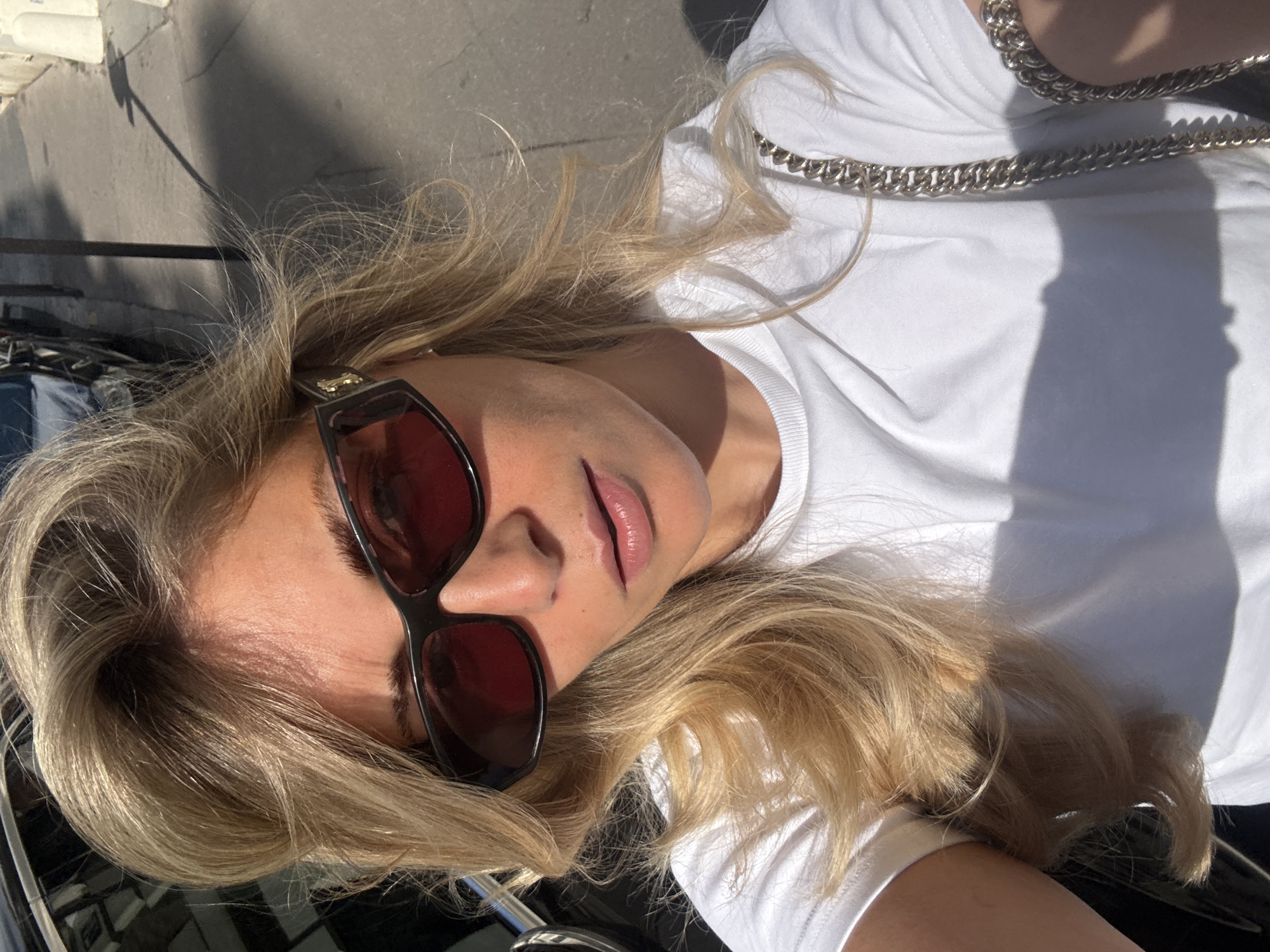 A blonde woman is lying on her back, wearing sunglasses, a white t-shirt, and a silver chain. The scene is outdoors, likely in Paris, France, given the style of architecture and lighting. A shadow is cast across her t-shirt and the background. She appears to be leaning against a black car.