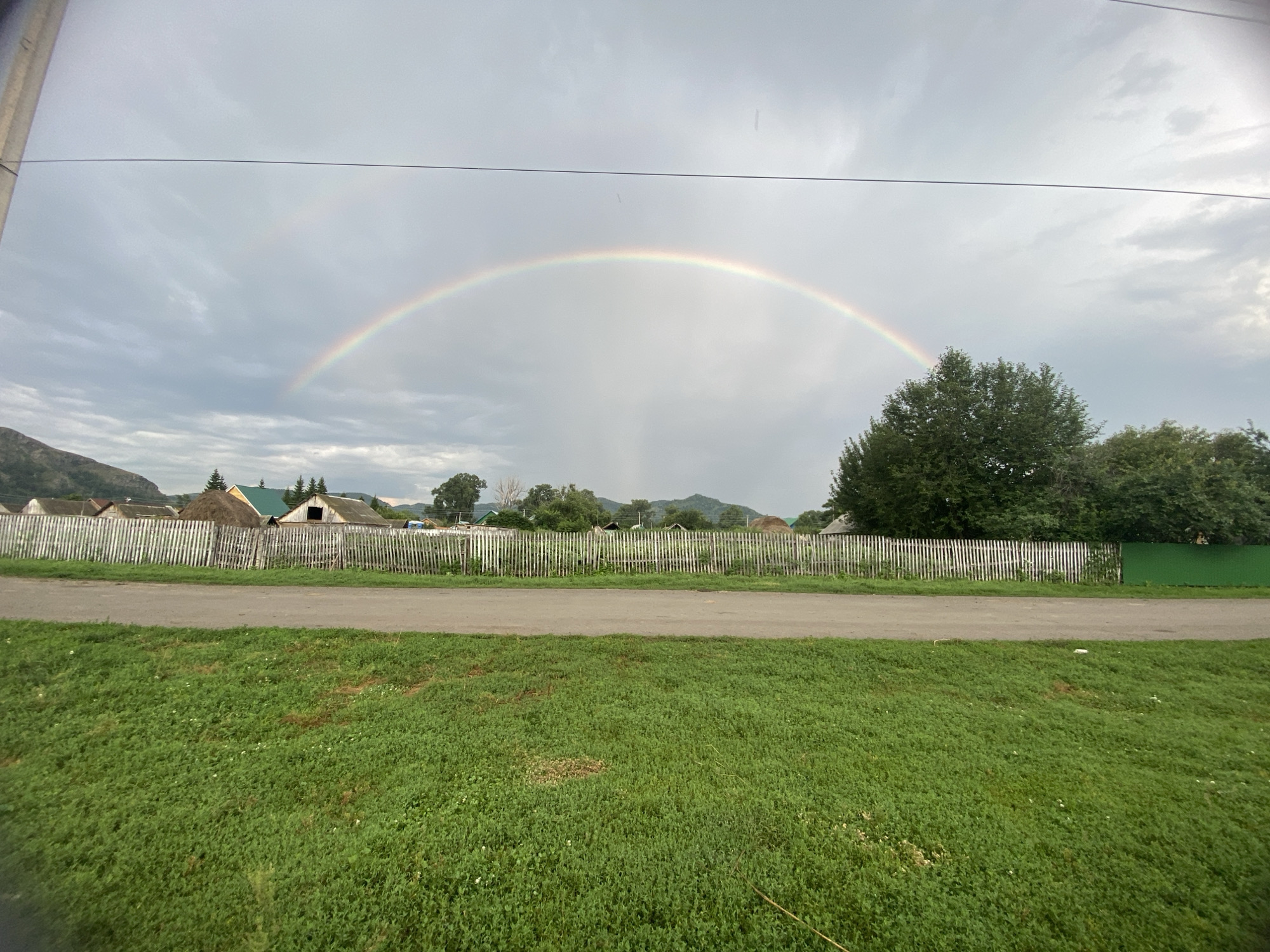 In Maloarslangulovo, Russia, a double rainbow arcs across a cloudy sky over a rural landscape. In the foreground, a strip of green grass leads to a dirt road. Beyond the road, a weathered white picket fence runs horizontally, separating the road from a village. Several rustic wooden houses with pitched roofs are visible behind the fence, some with haystacks. A large, lush green tree stands to the right of the frame, partially obscuring the village and a distant green fence. The overall scene depicts a tranquil, pastoral setting under a sky hinting at recent or impending rain.