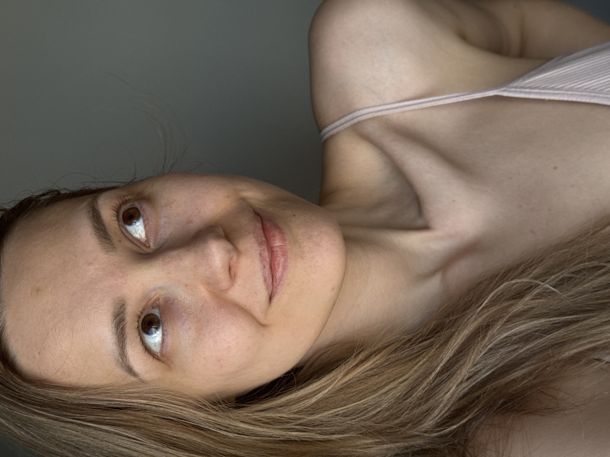 This close-up portrait features a young adult woman, depicted from the chest up and oriented sideways. She has fair skin, with visible freckles across her face and décolletage, and light brown hair that cascades towards the bottom of the frame. Her dark eyebrows frame expressive brown eyes, which are wide and directed upwards, reflecting a soft light source. A gentle, relaxed smile plays on her lips. She appears to be wearing a light-colored, possibly off-white or pale pink, striped camisole or slip, with a thin strap visible over her right shoulder and across her chest.
The background is a plain, solid medium-gray color, suggesting an indoor setting with minimal distractions. The lighting is soft and even, highlighting her features without harsh shadows, which might indicate natural light from a window or diffused artificial lighting. There are no specific objects or elements in the background that provide further context for the scene, beyond its indoor nature. No text is visible in the image. The overall impression is one of candid intimacy or quiet contemplation. The image was taken in Chaikovsky, Russia.