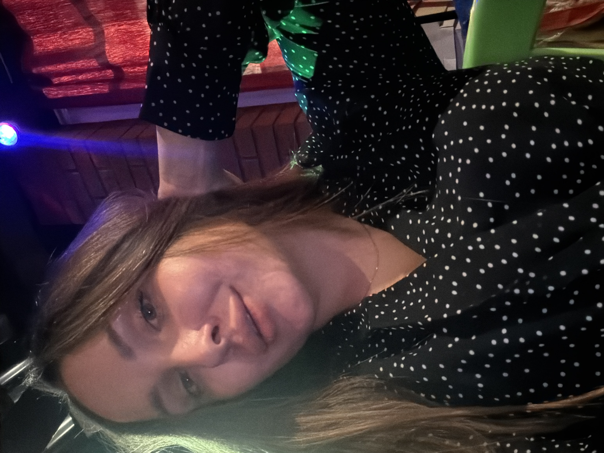 A young woman with long brown hair takes a selfie indoors. She is wearing a black dress with white polka dots and a thin gold necklace. Her left hand is resting on the back of her head. The background appears to be a dimly lit room with a brick wall, red curtains, and a blue light emanating from above. The woman's face is illuminated by green and pinkish light. The location is identified as Chaikovsky, Russia.