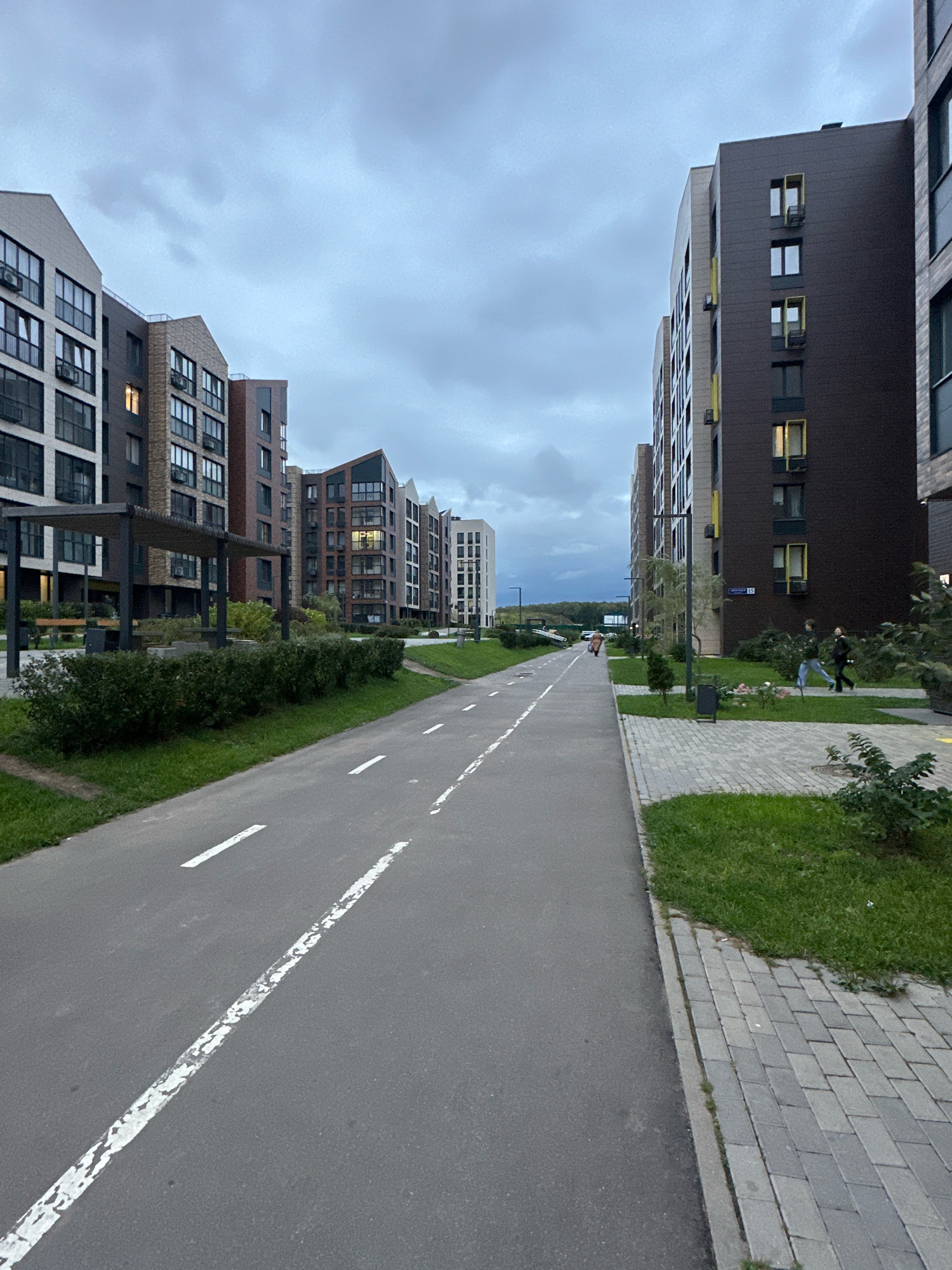 The image depicts a street scene in the city of Vidnoye, Russia, likely in a residential complex, as indicated by the modern apartment buildings lining both sides of a paved pathway. The pathway is marked with white lines, suggesting it serves as a pedestrian walkway or a cycling path. Green grass and landscaped bushes border the pathway. On the right side of the pathway, there is a sidewalk paved with gray interlocking bricks. Several apartment buildings are visible, characterized by their contemporary architectural style, featuring multiple stories, numerous windows, and varied facade colors, including brown, beige, and gray. In the middle ground of the image, two individuals are walking on the grass area between the pathway and the apartment buildings. The sky is overcast, with gray clouds. The overall atmosphere is calm and residential.