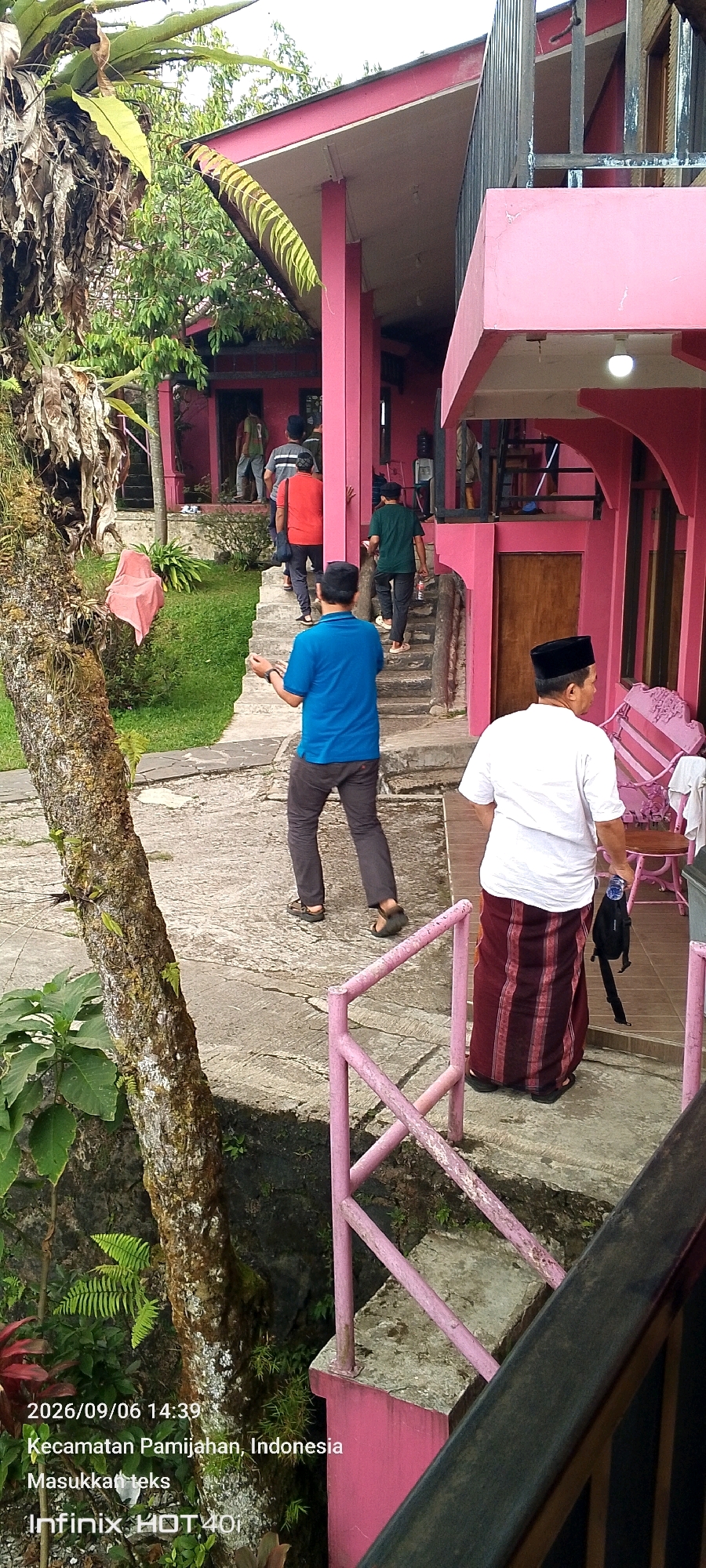 The image captures an outdoor scene in Pamijahan, Indonesia. Several individuals are present on concrete steps leading up to a pink building. On the left side of the steps, a person in a blue shirt and dark pants walks away from the camera. Further up the steps, a person in an orange shirt and dark pants ascends, followed by individuals in gray and green shirts. To the right of the steps, closer to the foreground, a person wearing a white shirt and a maroon and white striped sarong stands facing away from the camera. The environment includes lush greenery, a textured tree trunk on the left, and a pink railing along the side of the steps. The building has a pink facade and a dark railing on an upper level. A pink bench is visible on the right side of the image. The overall impression is a casual gathering or movement within the vicinity of the building.