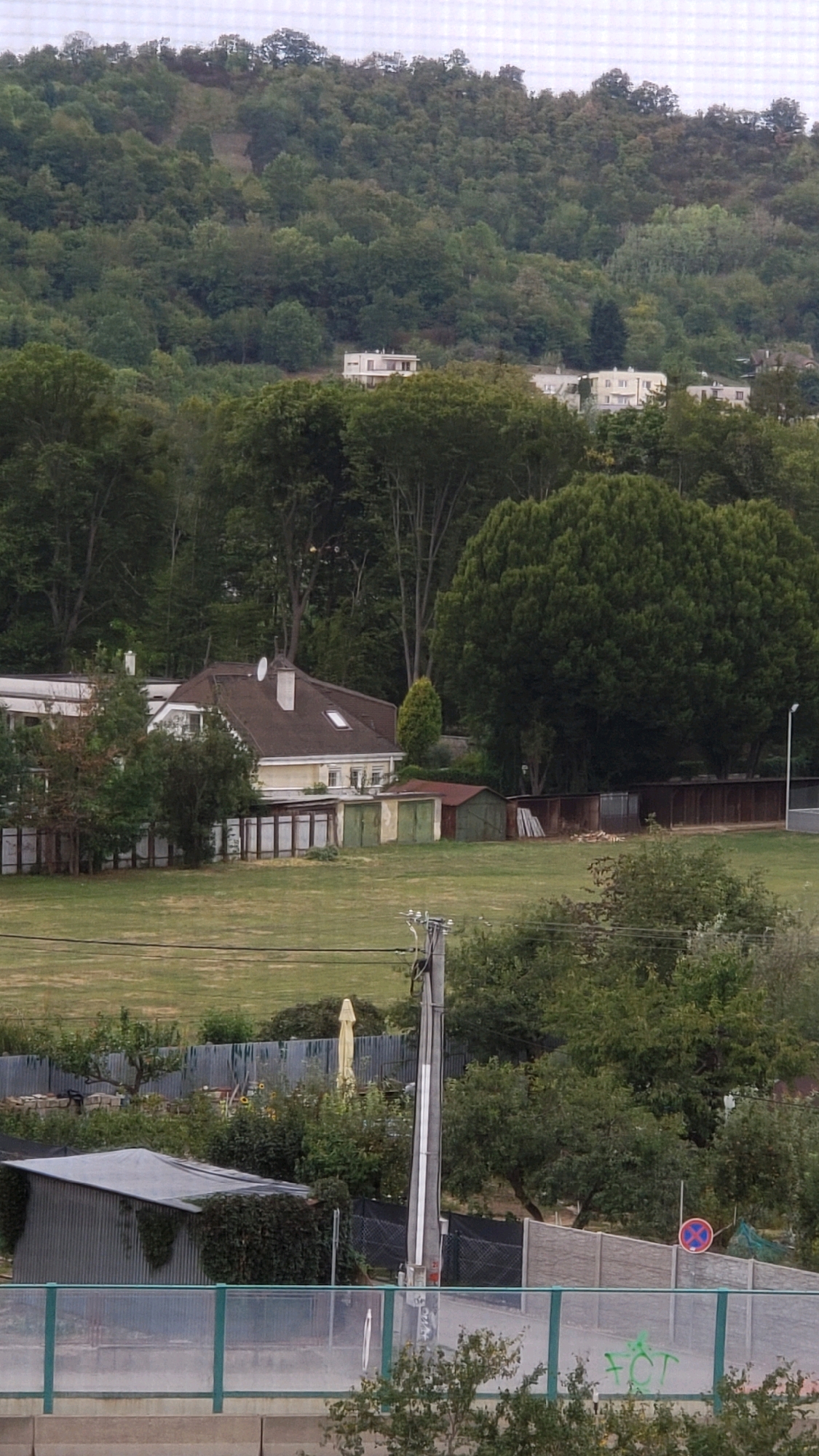 The image depicts a view of houses nestled among trees on a hillside. In the foreground, a grassy field is visible, bordered by a fence. Beyond the field, a house with a brown roof and white trim stands out. Several garages with green and brown doors are also present. The background features a dense forest covering the hillside, with glimpses of more houses in the distance. A utility pole with electrical wires runs through the scene. The overall environment suggests a semi-rural or suburban setting, possibly on the outskirts of a city.