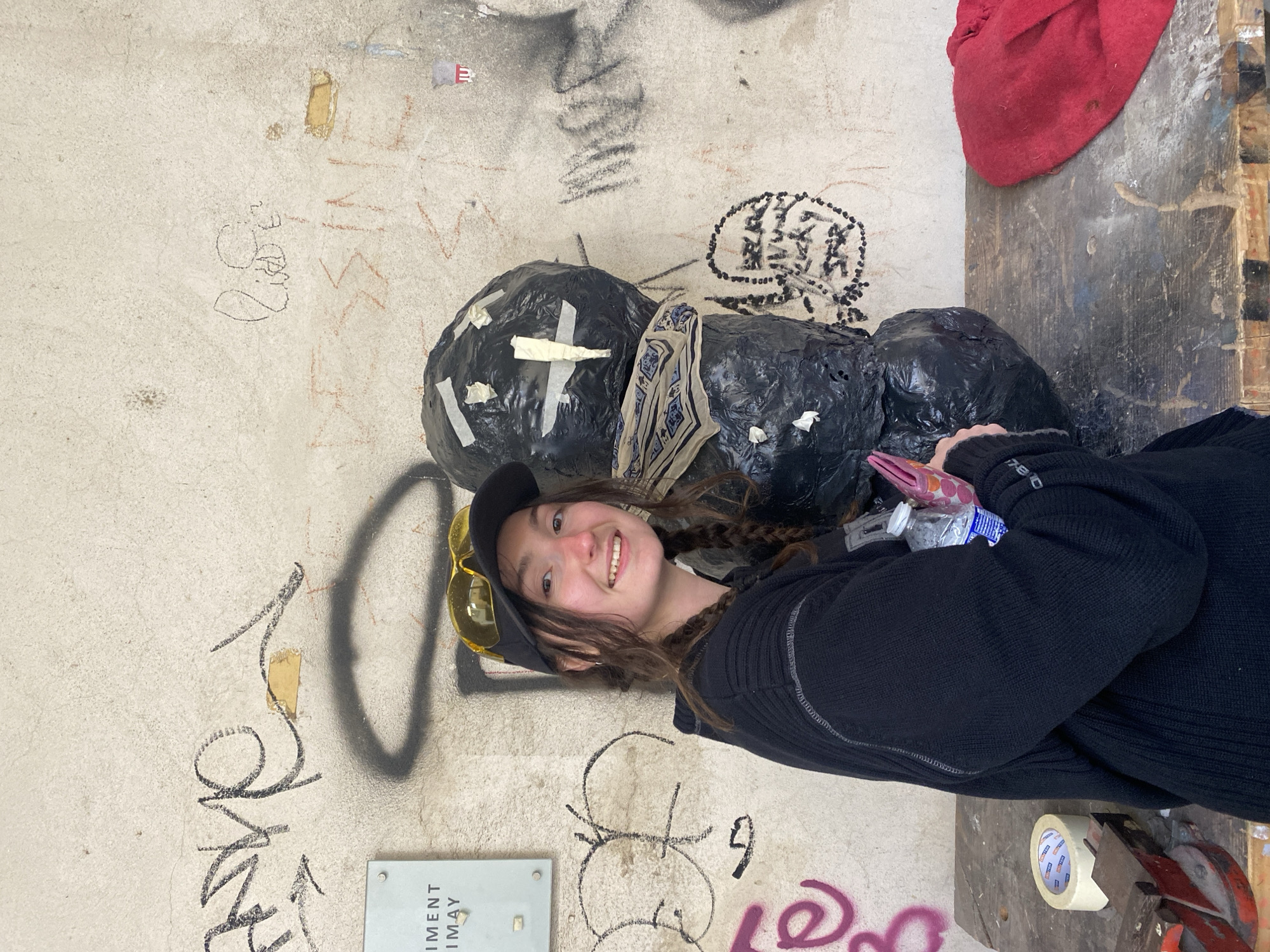 A young person with braided brown hair, wearing a black baseball cap and sunglasses, is smiling at the camera. They are wearing a black sweater and are holding a small wallet and a plastic bottle. They are standing next to a sculpture made of black trash bags, duct tape, and a patterned scarf. The sculpture is in front of a wall covered in graffiti. The wall has a small metal sign on it that reads "Experiment EIMAY." There is a table next to the person with a roll of tape and a tool. The photo was taken in Paris, France.