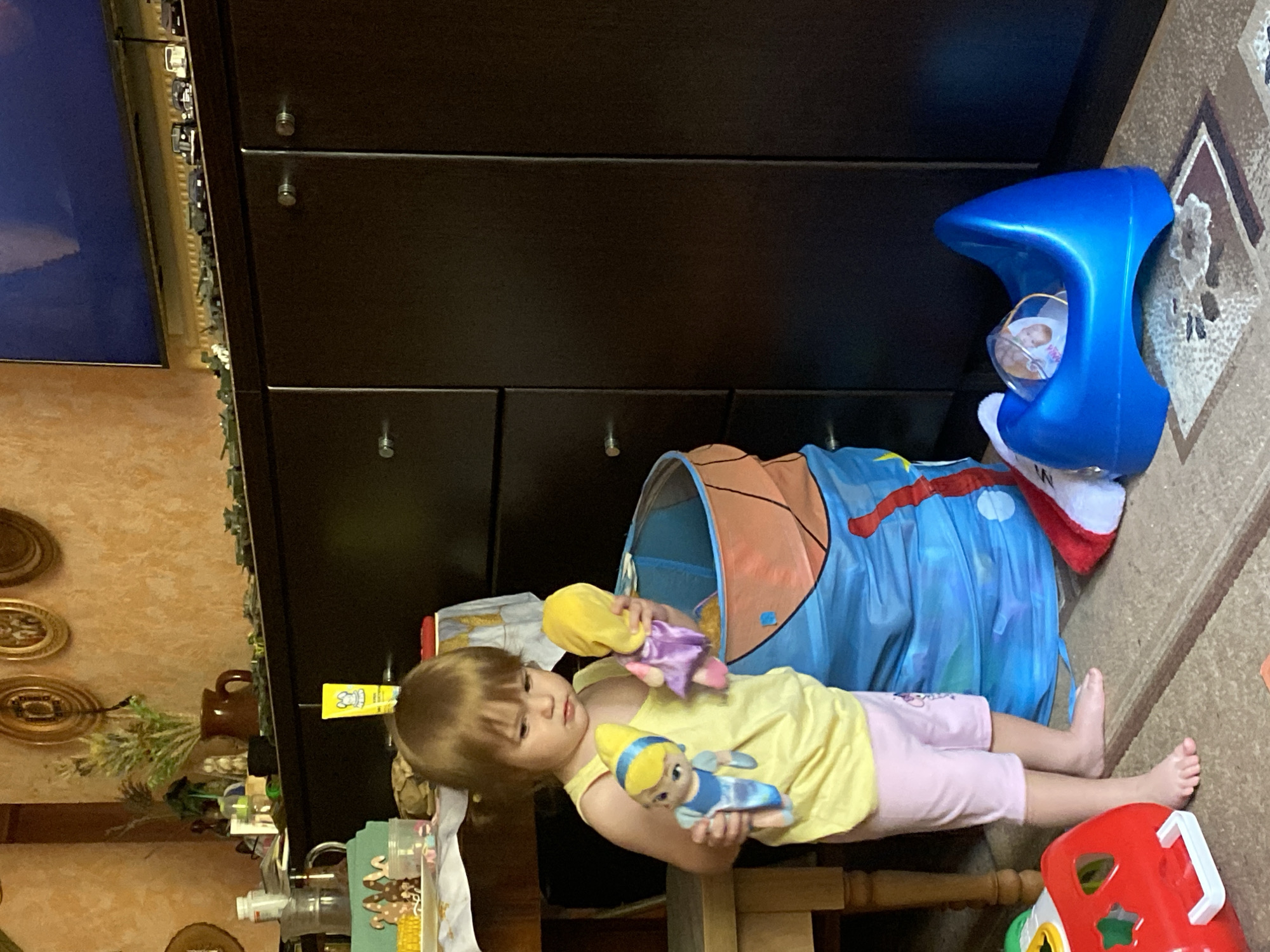 A young girl stands in a room, holding two dolls. She is wearing a yellow tank top and pink shorts. In front of her is a large blue toy bin. To her right is a blue potty training seat. A red toy is on the floor to her left. The room appears to be a living room or playroom, with a dark brown cabinet against the back wall. The walls are an orange textured material. There are various decorative items on shelves above the cabinet, including framed pictures and plants. Miniature toy vehicles are lined up on top of the cabinet.