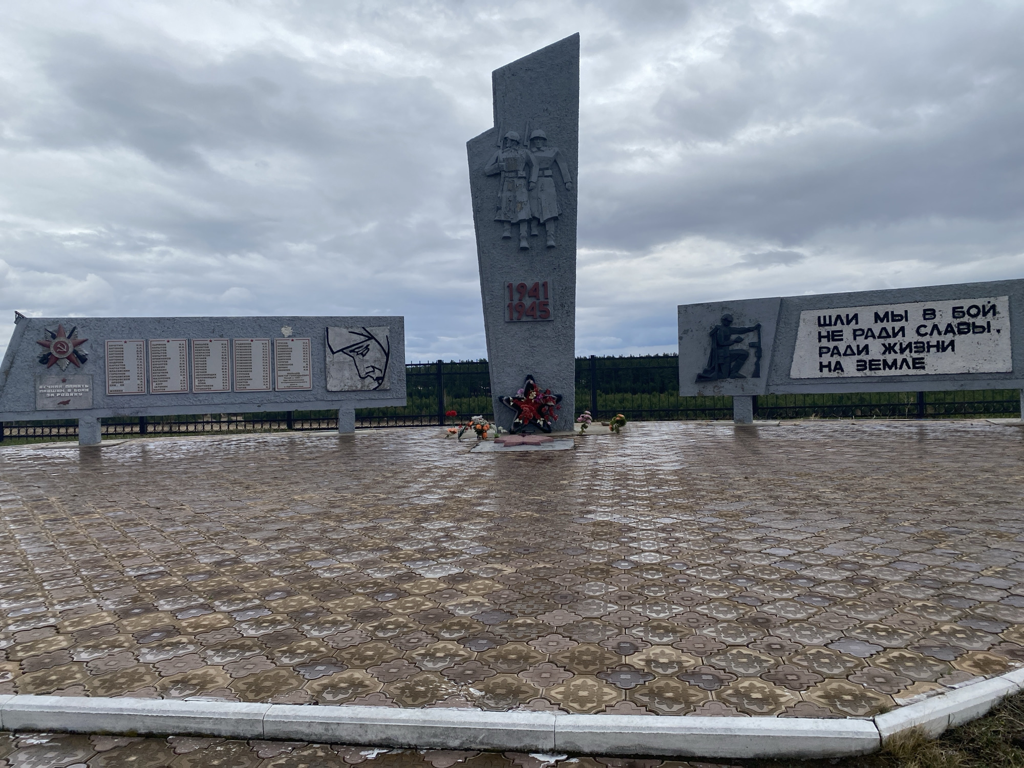 The image captures a memorial site dedicated to the soldiers of World War II, located in the city of Novomaltinsk, Russia. The scene is set on a cloudy day, indicated by the overcast sky. The ground is covered in patterned paving stones, which appear to be wet.
The memorial comprises three main concrete structures. The central structure features a relief of two soldiers and the dates "1941-1945" etched into the stone. To the left and right of the central structure are two rectangular concrete walls, each adorned with plaques. The left plaque displays a red star and a list of names, presumably of fallen soldiers. The right plaque has a figure of a soldier kneeling and a quote in Russian that translates to "We went into battle not for glory, but for life on earth."
At the foot of the central structure lies a collection of artificial flowers, laid in remembrance. The site is enclosed by a dark green metal fence, with trees visible in the background. The overall impression is one of solemn remembrance and respect for those who served during the war.