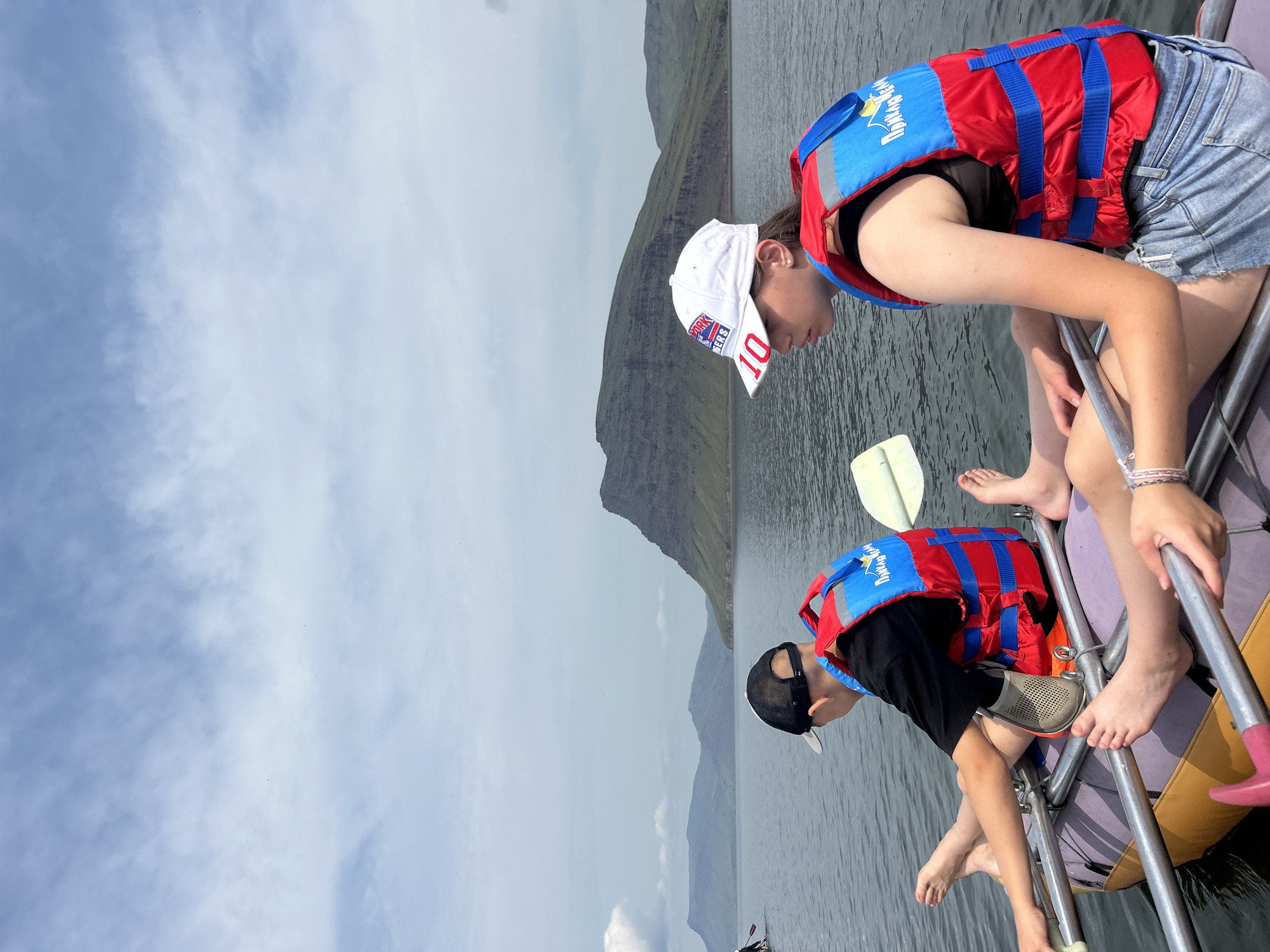 Two young people are in an inflatable raft on a body of water. The person on the left, wearing a black t-shirt and a black baseball cap, is looking away from the camera. They are wearing a red and blue life vest with some text on it. Their bare feet are visible. The person on the right, wearing a white baseball cap with "NEW YORK RANGERS" and the number "10" on it, is looking to the left. They are also wearing a red and blue life vest and denim shorts. Their hands are resting on the side of the raft, and their legs are visible. A paddle is resting across the raft. In the background, there are large, layered mountains under a cloudy sky. The scene appears to be in or near Nikolo-Petrovka, Russia, given the presence of the life vest text and the landscape.