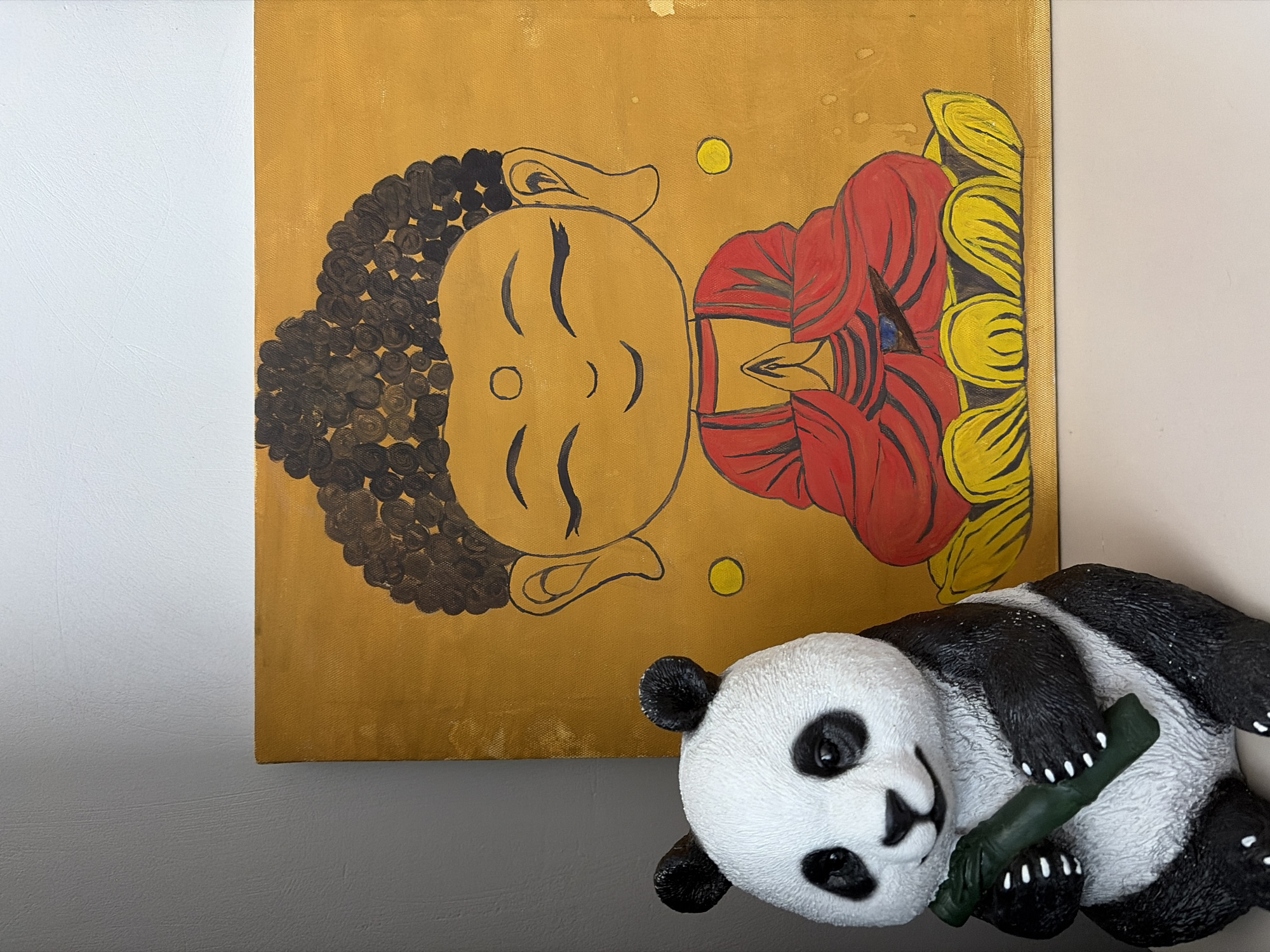A panda figurine and a painting of Buddha are positioned on a white surface, with a gray wall in the background. The panda, which is black and white, is holding a green bamboo stalk in its paws. The painting depicts a stylized, cartoon-like Buddha with closed eyes, wearing a red robe and sitting on a yellow lotus flower. The background of the painting is a vibrant golden yellow. The scene is well-lit, with the light source appearing to be from the front and slightly to the left, casting soft shadows. The overall impression is one of peaceful decoration, possibly in a home or a shop. The location in Kyzyl, Russia is not discernible from the visual information provided in the image.