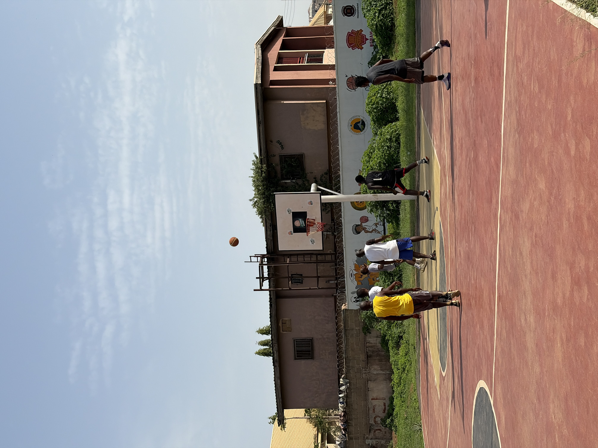 The scene is an outdoor basketball court in Gura Top, Nigeria. Several men play basketball, with one shooting at the hoop. The court is red with white lines and painted arcs. Behind the hoop are buildings, one with a mural-decorated wall, bordered by green shrubbery. The sky is blue with some clouds.