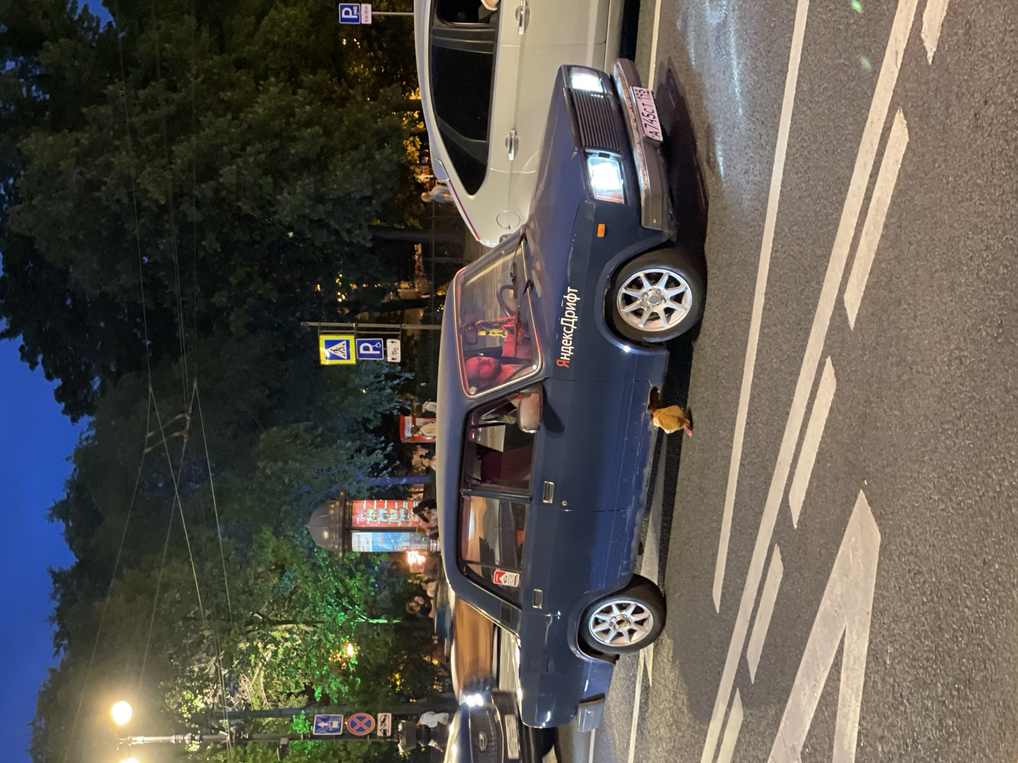 This image captures a nighttime street scene in Saint Petersburg, Russia, showing multiple vehicles and pedestrians under artificial lighting.
In the foreground, a dark blue, older model Lada sedan, bearing the Russian license plate A745CT 198, is stopped in the right lane. The car features custom white multi-spoke wheels and a white "ЯндексДрифт" (Yandex Drift) decal on the front passenger side of the hood. A "Marlboro" sticker is affixed to the rear passenger window. A male driver, wearing a red top, is visible through the windshield, with his hands on the steering wheel. A small yellow/orange object, possibly a toy, hangs below the front bumper on the right side. The car's headlights are illuminated, casting light onto the asphalt road.
To the immediate right of the blue sedan, a white hatchback (appears to be a Ford Focus) is also stopped, partially visible. To the left, a dark-colored vehicle is partially visible. In the background, a blurred, light brown/orange vehicle indicates motion.
The road surface is asphalt, marked with distinct white dashed and solid lines, consistent with a pedestrian crossing area. Numerous traffic signs are present on poles along the street. On the left, signs include a pedestrian crossing indicator, a 'No Stopping' sign, and a 'Parking' sign. On the right, signs include a pedestrian crossing warning triangle and 'Parking' signs with supplementary information.
The background features dense green foliage from numerous trees, some illuminated with a green hue by streetlights. The sky is a deep dark blue. Overhead wires, likely for trams or trolleybuses, traverse the upper portion of the frame. A prominent cylindrical advertising column with a dark domed top, displaying various illuminated advertisements, stands in the mid-ground. Behind the white car, a red structure resembling a telephone booth is visible. Several pedestrians are scattered in the mid-ground, some near the advertising column and others further back, appearing to be standing or walking. A bright lamppost with two illuminated yellow lights is visible on the far left.