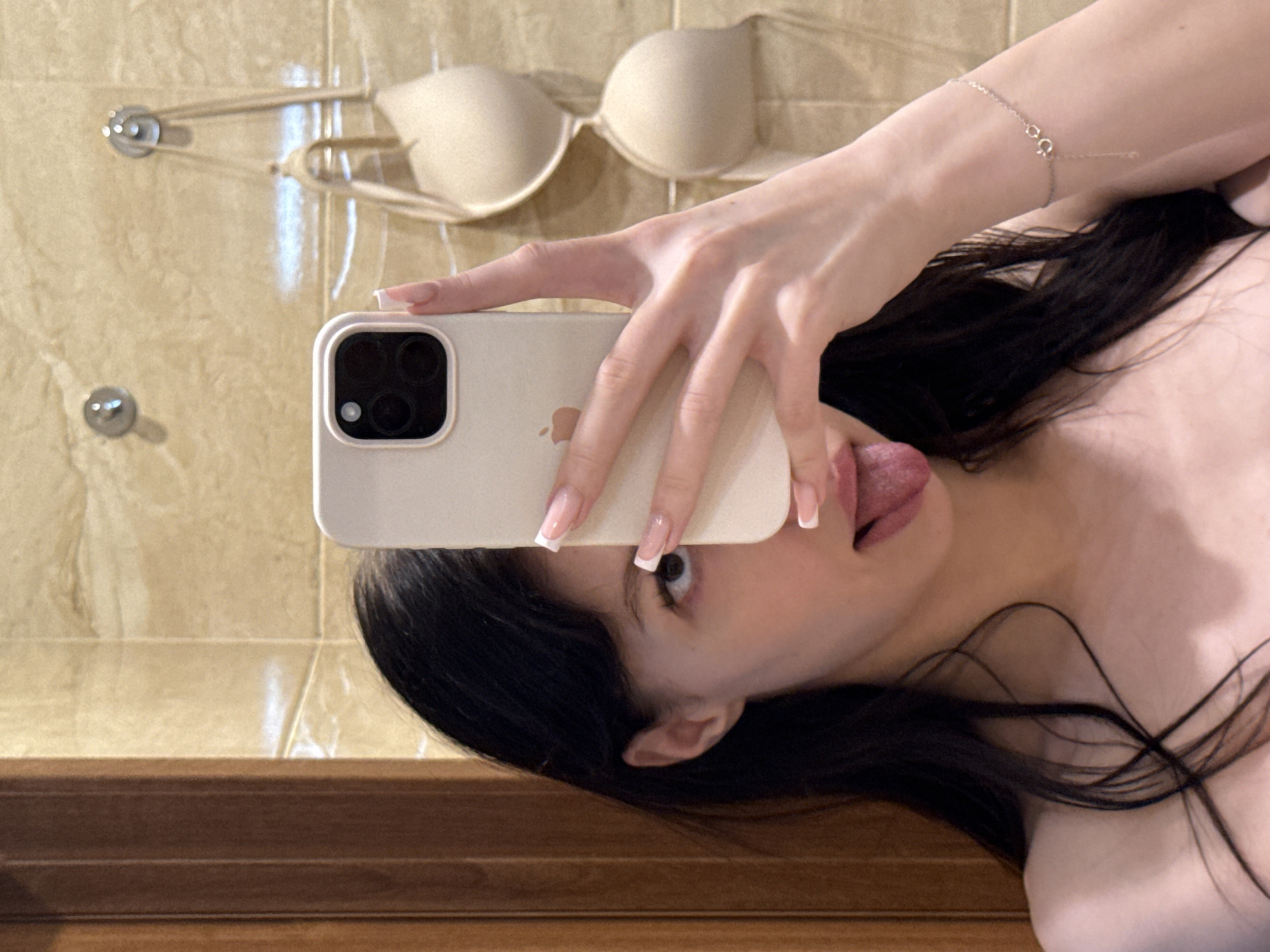 A young woman with long, dark hair is taking a selfie in a bathroom. She is holding a white iPhone in her right hand, obscuring the right side of her face, with her tongue sticking out. She is wearing a thin-strapped bracelet on her left wrist. In the background, a beige bra is hanging on a hook on a tiled wall. The wall to the left of the image appears to be made of wood. There are no identifiable landmarks that place the image specifically in Khimki, Russia.