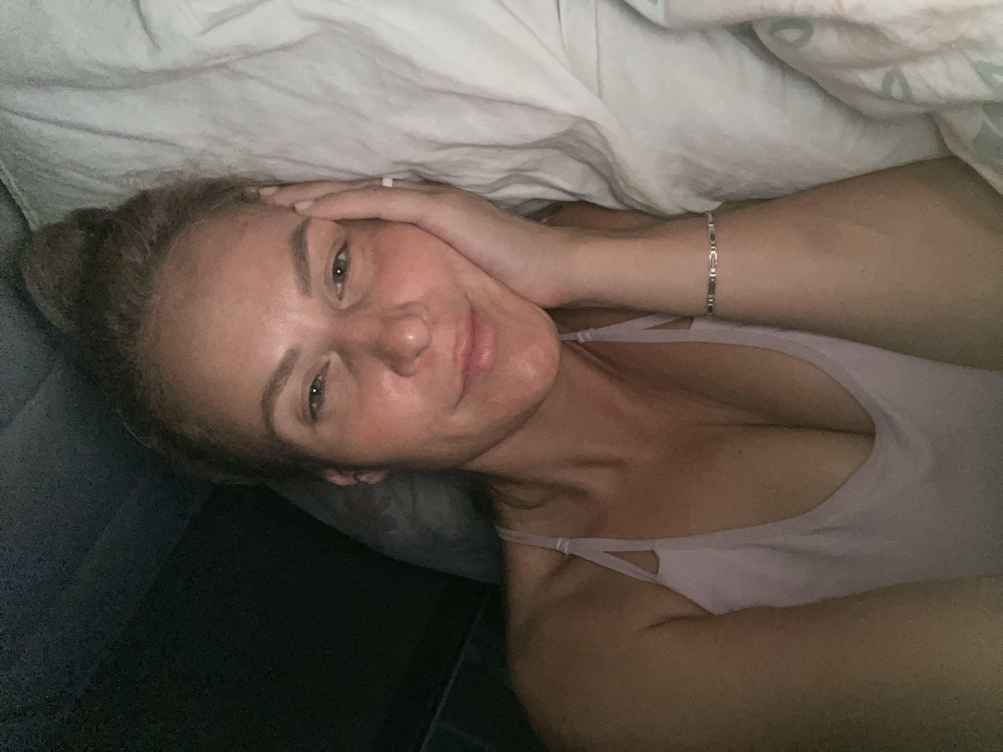 A woman is lying in bed, partially under a white blanket. She is wearing a light pink tank top and a silver bracelet on her left wrist. Her left hand is touching her face. The location of the city of Baranovka, Russia, is not discernible from the image.