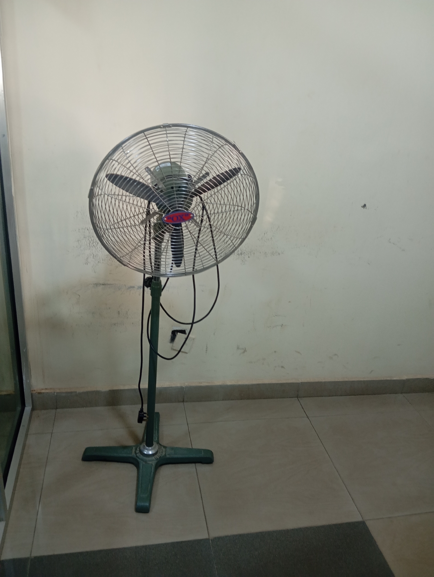 An indoor scene features a tall, oscillating fan with a silver metal grille and dark blades, standing on a dark green, cross-shaped base. The fan is positioned in front of a light-colored wall with visible smudges and marks. The floor is tiled with large, square, light brown tiles, with a darker tiled area to the lower right. The fan's black power cord hangs down its green stand. This scene is located in a room in the city of Jos, Nigeria.