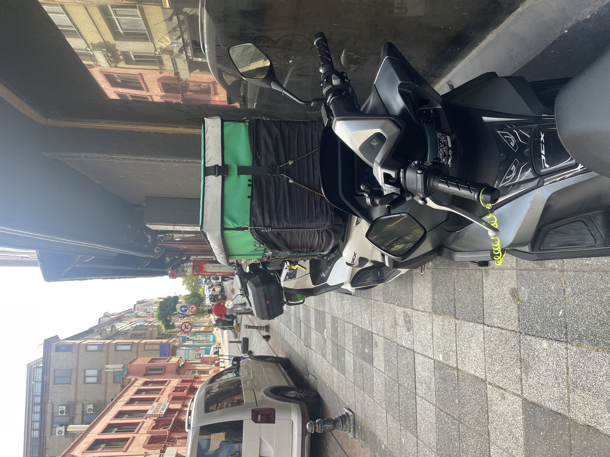 An eye-level, medium shot captures a delivery scooter parked on a cobblestone sidewalk on a street in Kadıköy, Turkey. The scooter, predominantly white and black, faces right and is equipped with a large, green and white insulated delivery bag strapped to its rear. A yellow cable lock is attached to the scooter's handlebars. To the left of the scooter, a white van is parked parallel to the curb. The street is lined with multi-story buildings, their facades painted in various colors including pink and beige. Several traffic signs, including a "no entry" sign and directional arrows, are visible further down the street. A person in a red shirt walks away from the camera towards the buildings. The sky is bright and overcast, indicating daytime.
