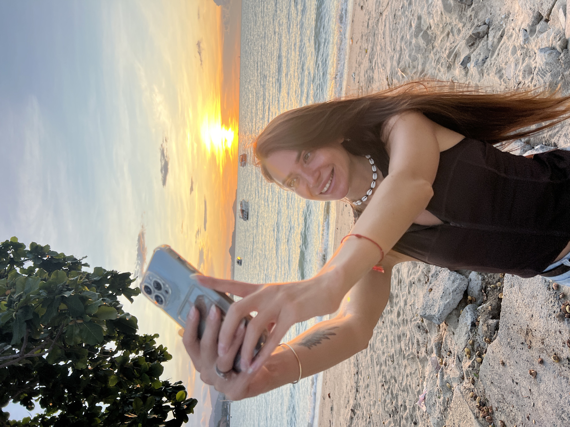 A woman is taking a selfie on a beach with the ocean and a sunset in the background. She is wearing a black top, a shell necklace, and has long brown hair. Her left arm is extended, holding up a smartphone. A tree branch with green leaves is visible in the upper left corner. Boats are scattered across the water. The sand on the beach has some rocks and debris. The sky is a blend of blue and orange from the setting sun.