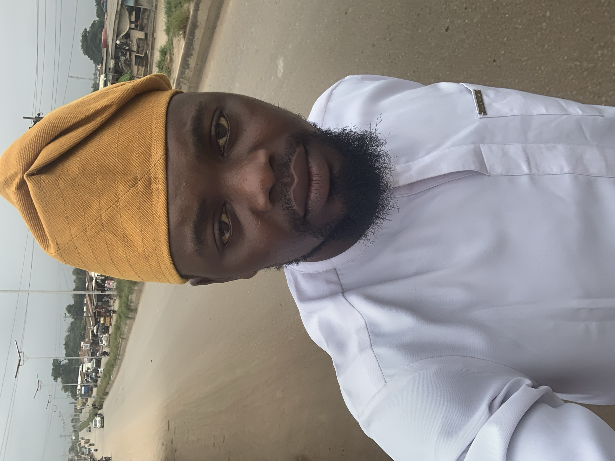 A man wearing a yellow cap and white traditional attire stands on a dirt road in Shagamu, Nigeria. The man is looking directly at the camera. In the background, there is a busy road with cars and motorcycles, as well as shops and buildings lining the street. The sky is overcast, and the overall impression is one of a typical day in a Nigerian city.