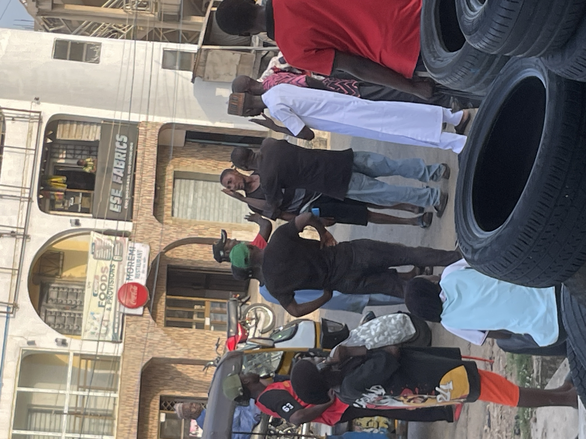 The image depicts an outdoor scene in what appears to be a commercial area. In the background, a multi-story building features signage for "FOODS & PROVISIONS" and "MOREMI RESTAURANT," as well as "ESE FABRICS." Several individuals are present in the foreground and midground. Some are standing, while others appear to be seated. A three-wheeled vehicle, possibly an auto-rickshaw, is parked on the left side of the frame. There are also several car tires stacked in the lower right portion of the image. The people are dressed in casual clothing, and some appear to be engaged in conversation or interaction. The overall atmosphere suggests a street scene in a bustling urban environment.