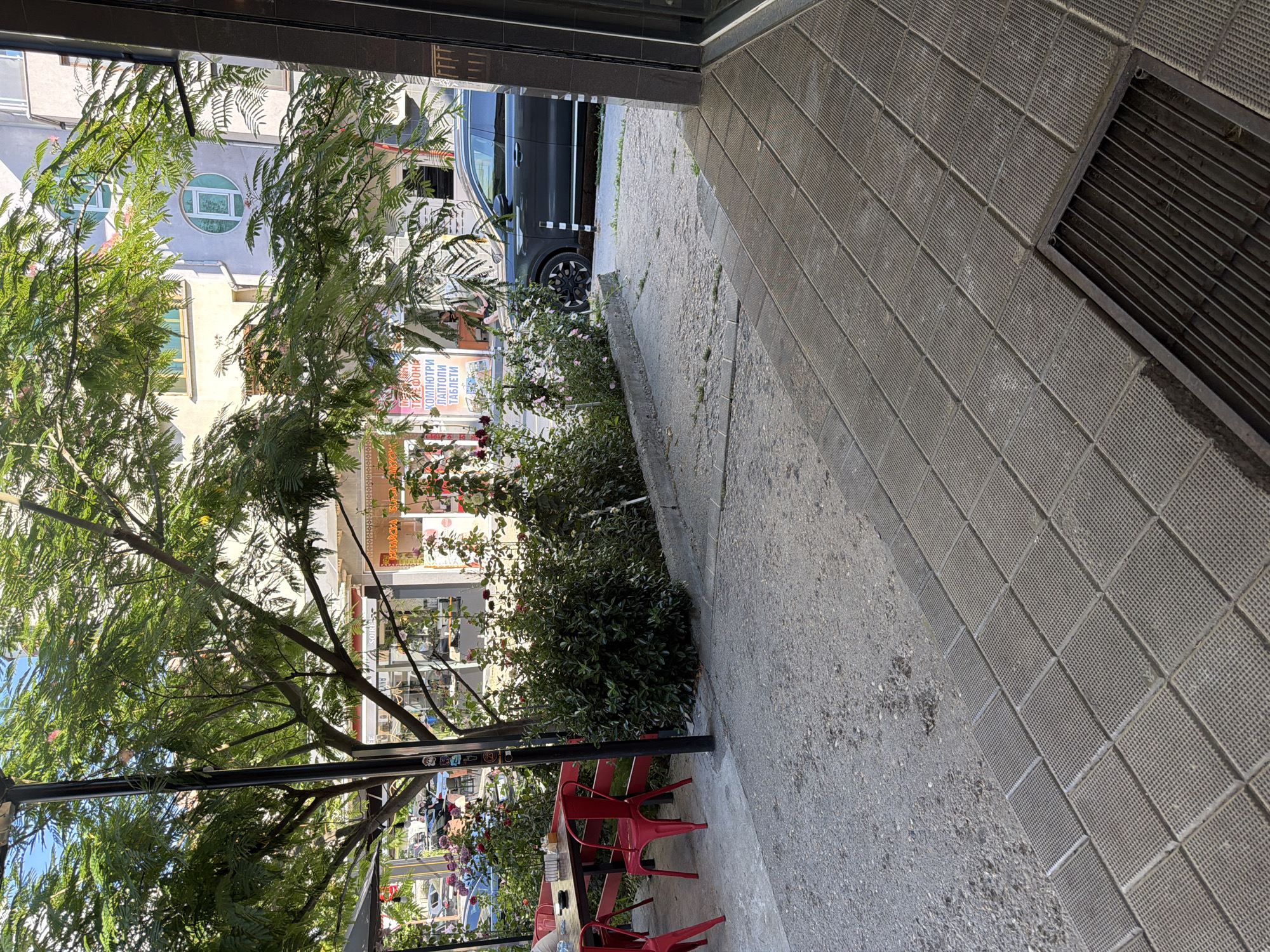 In Basarbovo, Bulgaria, a sidewalk cafe area is depicted. A dark-colored SUV is parked on the street adjacent to a walkway. Several red chairs and a wooden table are set up under the shade of a large tree with green foliage, suggesting an outdoor dining or seating area. Behind the cafe setup, a building with windows, some circular, is visible, indicating an urban environment. The foreground shows a textured concrete sidewalk with a grate.