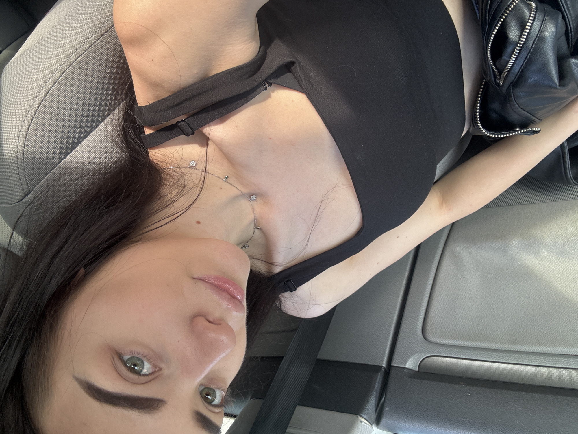 A selfie captures a woman in what appears to be a car interior. She is wearing a black top and a delicate necklace. A black leather jacket is visible next to her. The seat of the car is gray. The location is unknown.