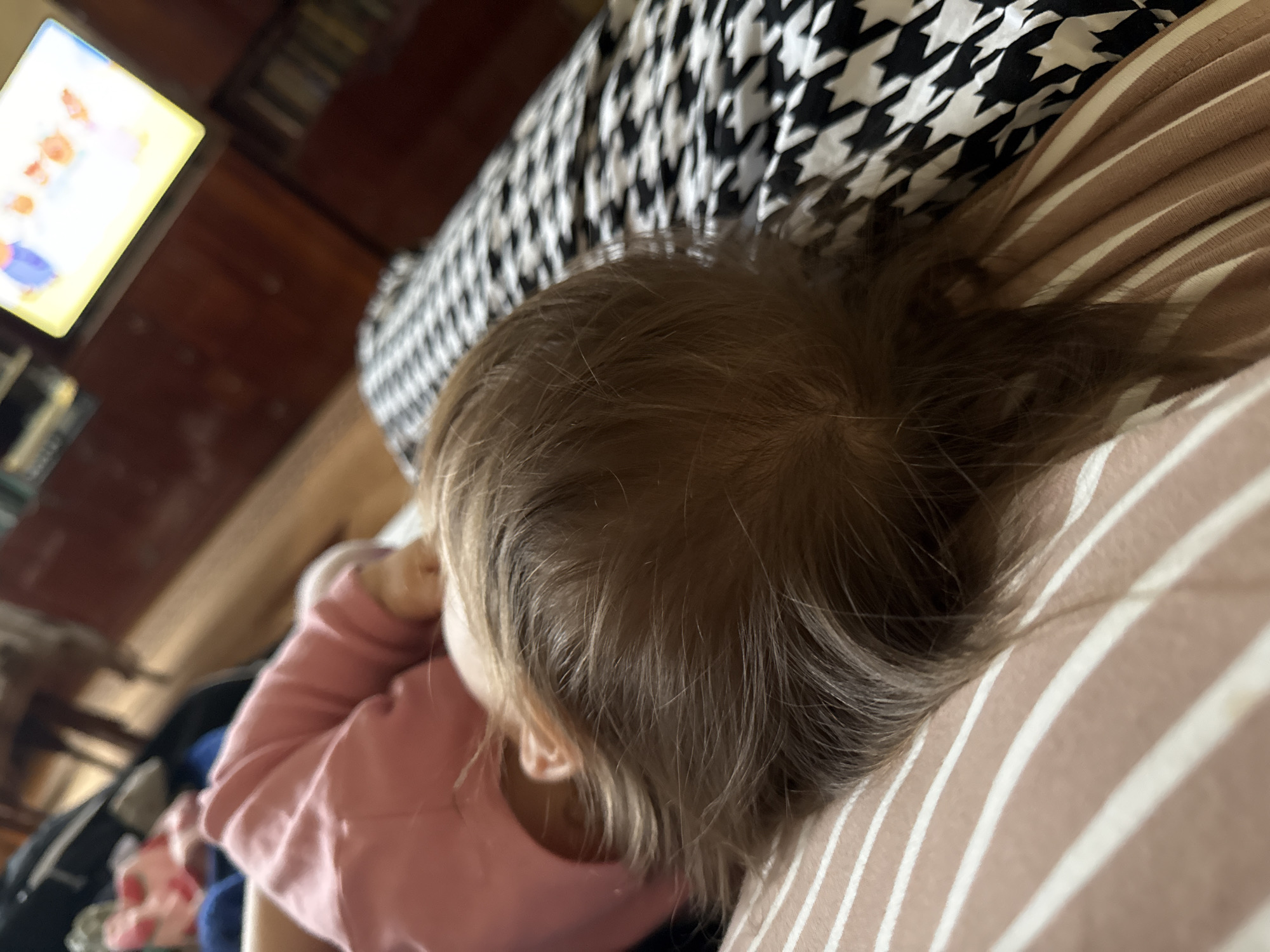 An overhead, slightly angled photograph captures a young child resting indoors, likely in a living room, in Krapivka, Russia. The child, visible from the top of their head and upper back, has light brown hair and is wearing a long-sleeved pink top. They are positioned on or next to a striped surface, which is light brown or beige with prominent white stripes. Behind the child, a black and white fabric featuring a houndstooth pattern is visible, suggesting a blanket or pillow.
In the blurred background to the left, a brightly lit rectangular screen, presumably a television or tablet, displays colorful imagery that appears to be a cartoon or show. Dark wooden furniture or paneling can be seen further back. The child's posture, with a bent arm, suggests a state of relaxation, possibly while watching the screen or simply resting. The scene conveys a calm, domestic atmosphere. No visible text is present in the image. The time of day and external weather conditions are not discernible from the indoor setting.