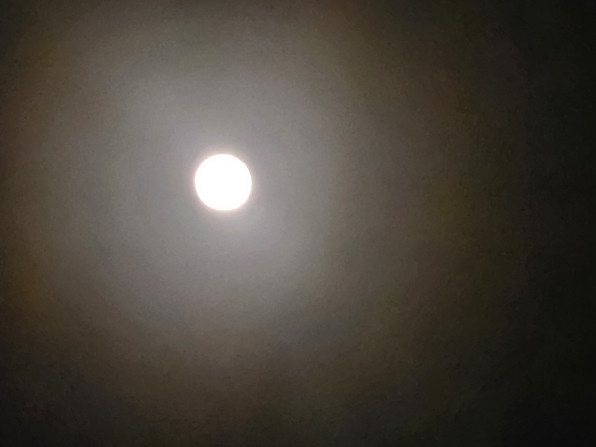 The image depicts a bright, circular light source, likely a lightbulb, positioned centrally in the frame. The light emanates outwards, creating a halo effect against a dark, textured background that appears to be a ceiling. The halo is somewhat diffused, suggesting a slight haze or dust in the air. There are subtle variations in the background's tone, transitioning from a lighter gray around the light source to a darker, almost black hue at the edges of the image. The overall impression is one of an indoor environment, possibly a room or an open space, illuminated by a single overhead light. The image does not contain any people, animals, or specific details that would pinpoint its location within Beer Tuvia Regional Council, Israel, or any other specific urban or rural setting.