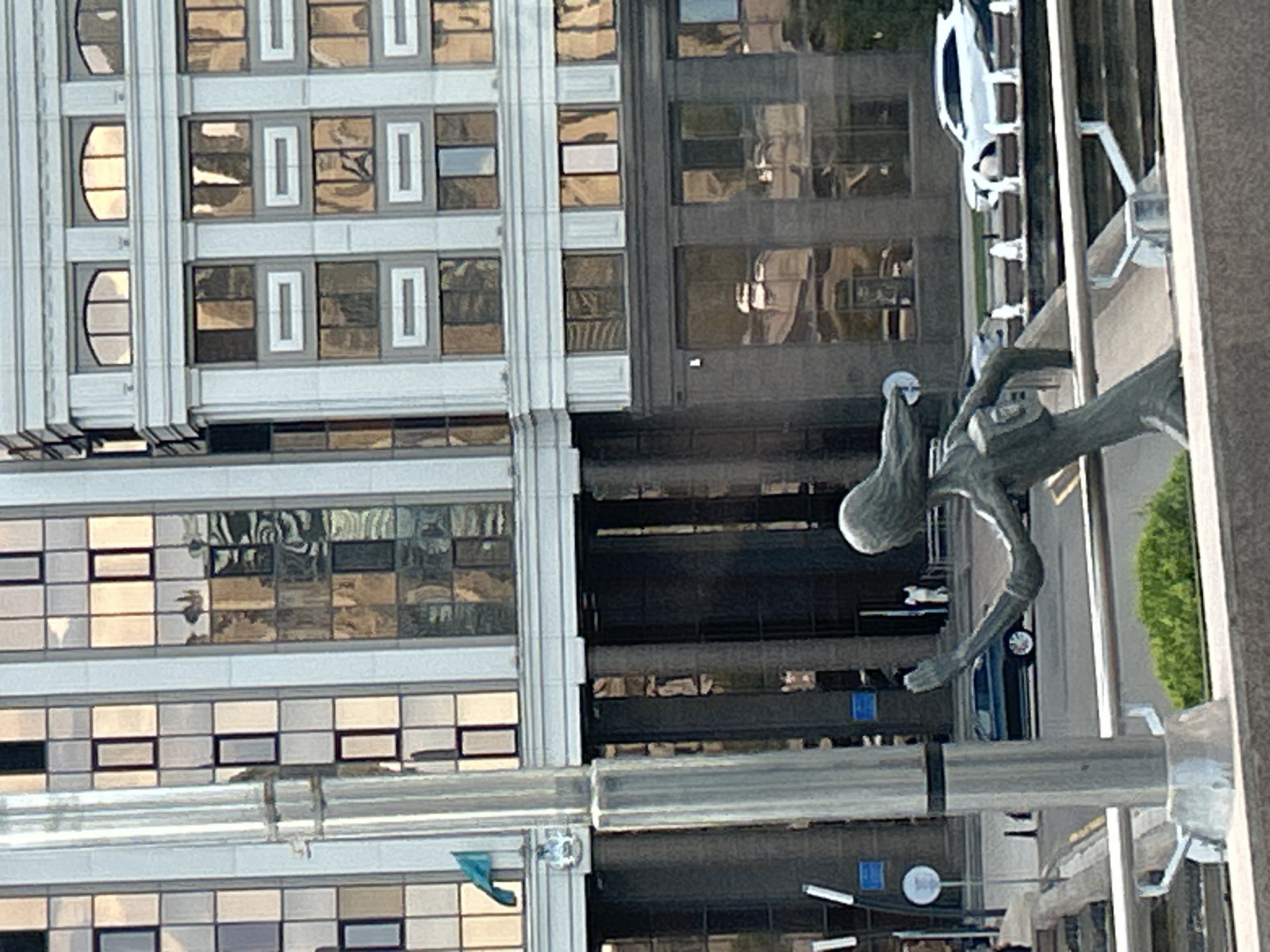 A bronze statue of a woman with flowing hair and outstretched arms stands in the foreground, facing right. She is positioned in front of a modern building with a facade of reflective windows and white architectural elements. The statue is partially obscured by a tall, cylindrical metal pole on its left. Behind the statue, a street is visible, with a dark-colored car driving on it and a white SUV parked further down. A fountain with multiple spouts is also present. The building in the background displays reflections of the sky and surrounding environment in its windows. A small green flag with a symbol is mounted on the building above the statue. The overall setting appears to be an urban plaza or public space in Astana, Kazakhstan.
