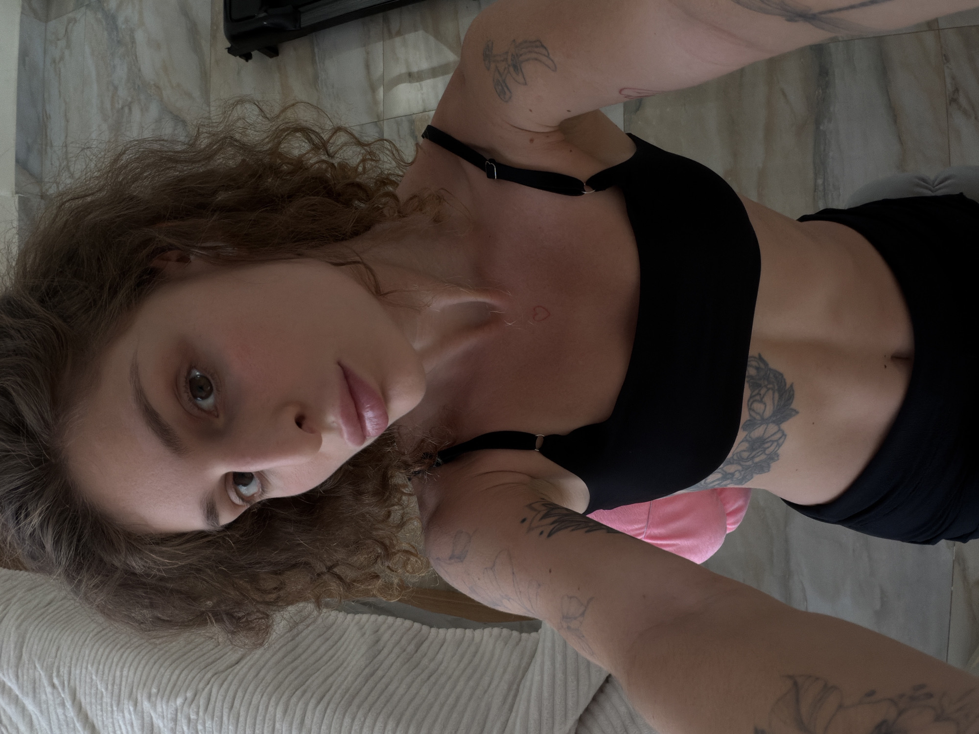 A woman with curly brown hair, light brown eyes, and full lips is taking a selfie indoors. She is wearing a black sports bra and black bottoms. Tattoos are visible on her arms and abdomen. A pink object, possibly a pillow or bag, is behind her. The flooring appears to be marble. A treadmill is partially visible in the background to the right. The background also includes a textured white surface, possibly a bed or couch, on the left. The scene is lit by natural light. The specific location in North Kuta, Indonesia, cannot be determined from the visual information alone.