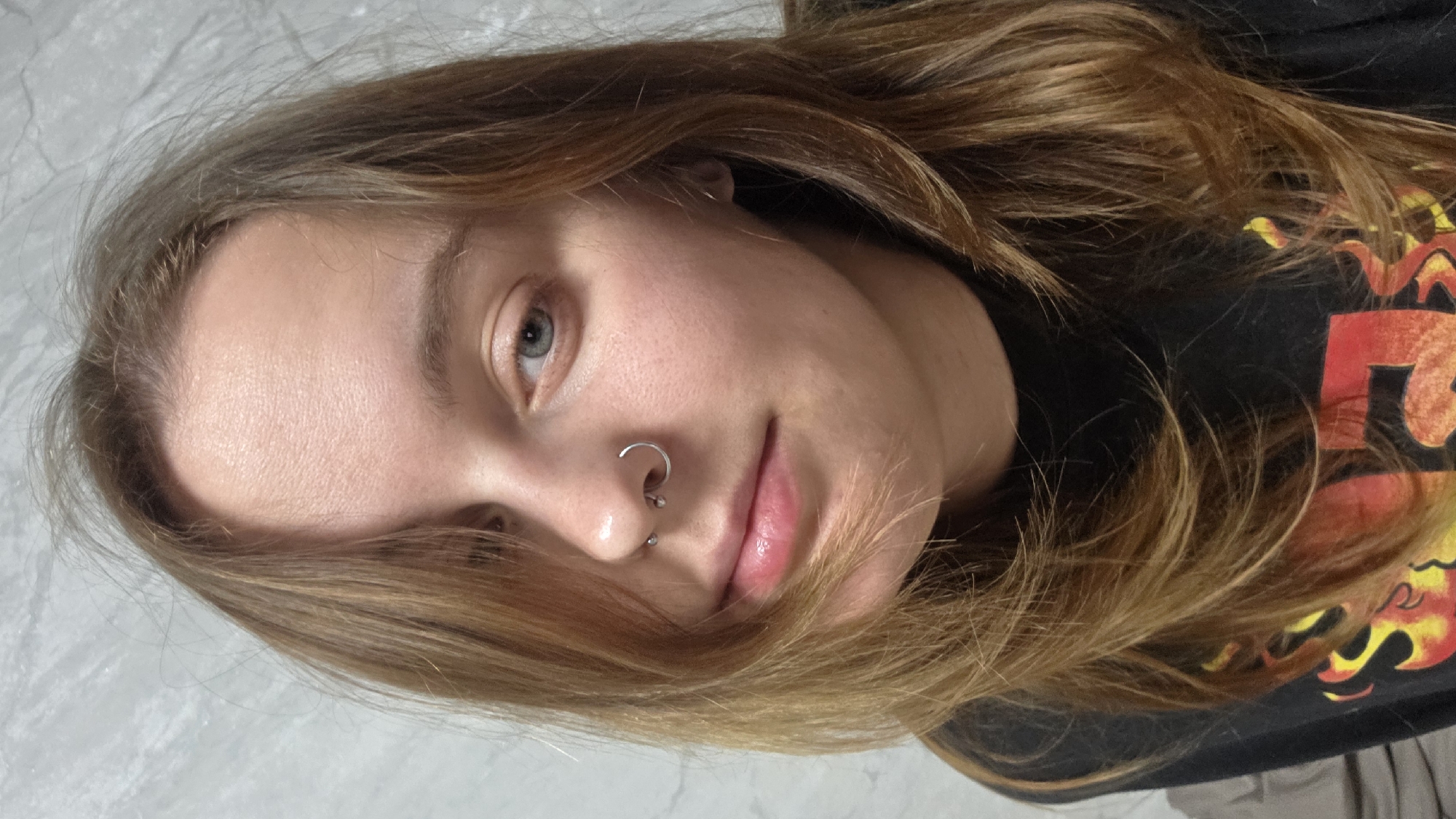 A person with fair skin and light brown hair is depicted in a close-up shot. The subject has light-colored eyes and facial piercings on the nose, including a ring and a stud. Some strands of hair fall across the person's face. The person is wearing a black t-shirt with a design in red and yellow. The background is a textured white surface. The setting or city is not identifiable from the provided image.