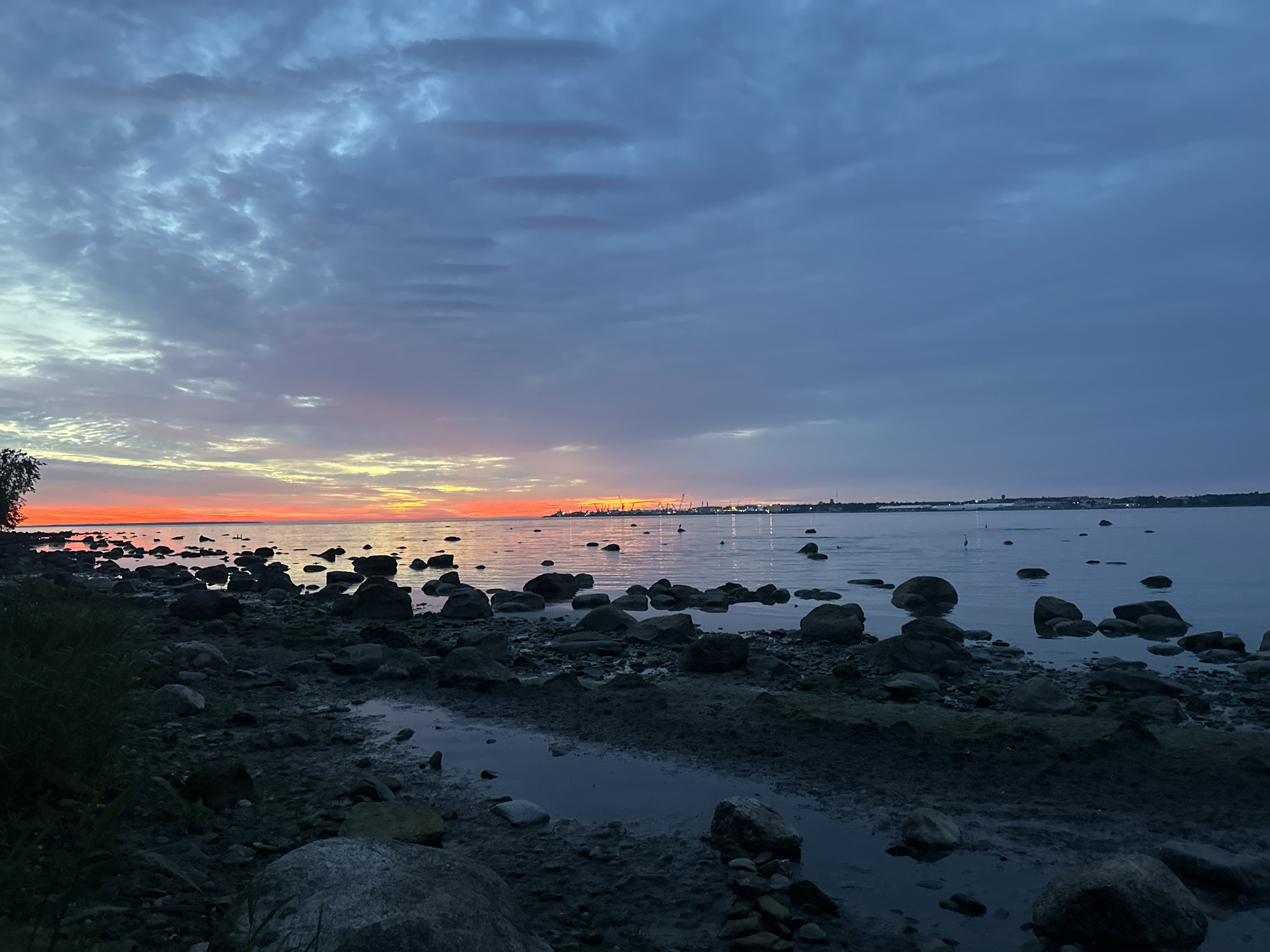 The image depicts a rocky shoreline along the Baltic Sea, likely in Tallinn, Estonia, during twilight. The sky is filled with dramatic clouds, tinged with hues of orange, yellow, and blue, indicating the setting sun. The water is calm, reflecting the colors of the sky. Numerous rocks of various sizes are scattered along the shore and in the water. In the distance, the city skyline of Tallinn is visible, with cranes and lights suggesting an industrial or harbor area. There are no visible people in the image. The overall scene is serene and captures the beauty of the natural landscape against an urban backdrop.