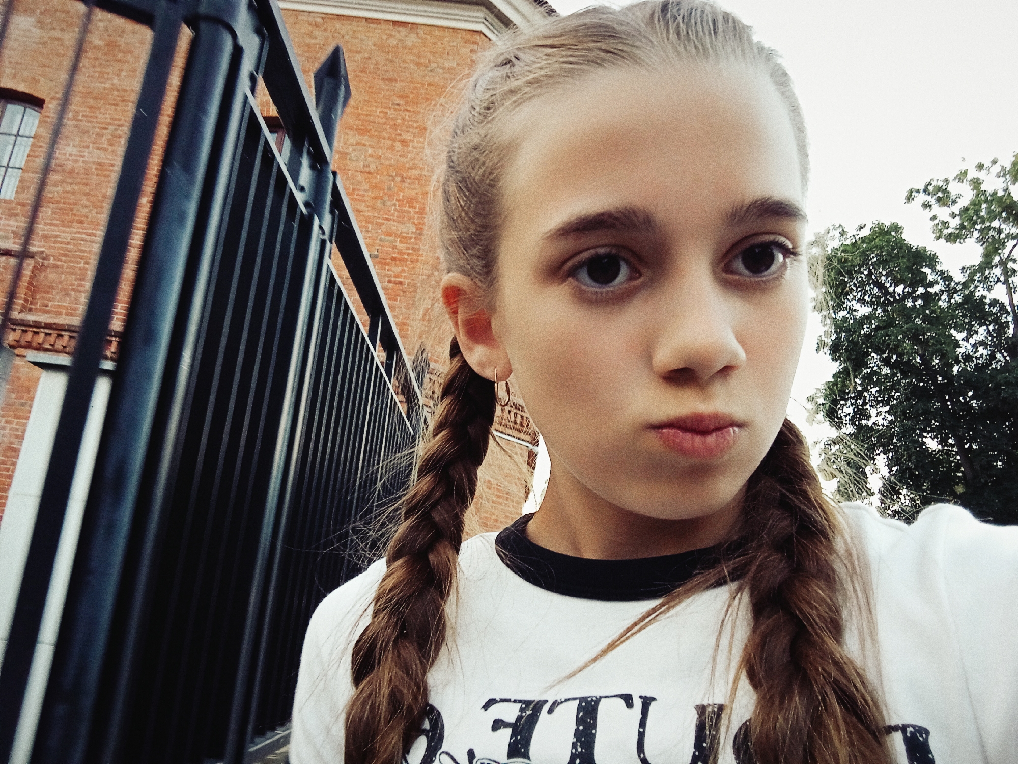 A young girl with braided hair is pictured, looking directly at the camera. She is wearing a white t-shirt with a black collar. Her face is partially obscured by the angle of the photograph. Behind her, a dark metal fence runs vertically, with a red brick building visible in the background. To the right of the frame, green foliage from trees is present against a bright sky. The setting appears to be outdoors, in an urban environment, possibly a park or the grounds of a historical building. There is no clear indication of the specific location within Warsaw, Poland.